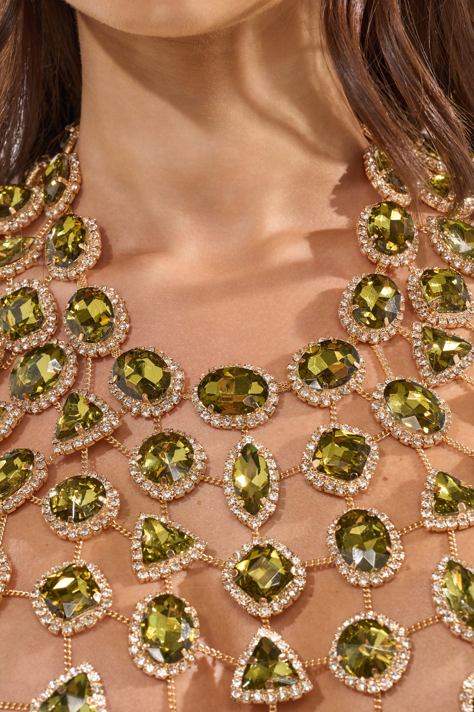 A close-up of a woman wearing the ALL I NEED IS MONEY RHINESTONE CHAIN TOP, featuring elaborate gold-colored metal with large green faceted gemstones and small clear stones draped elegantly across her chest and neck. 8