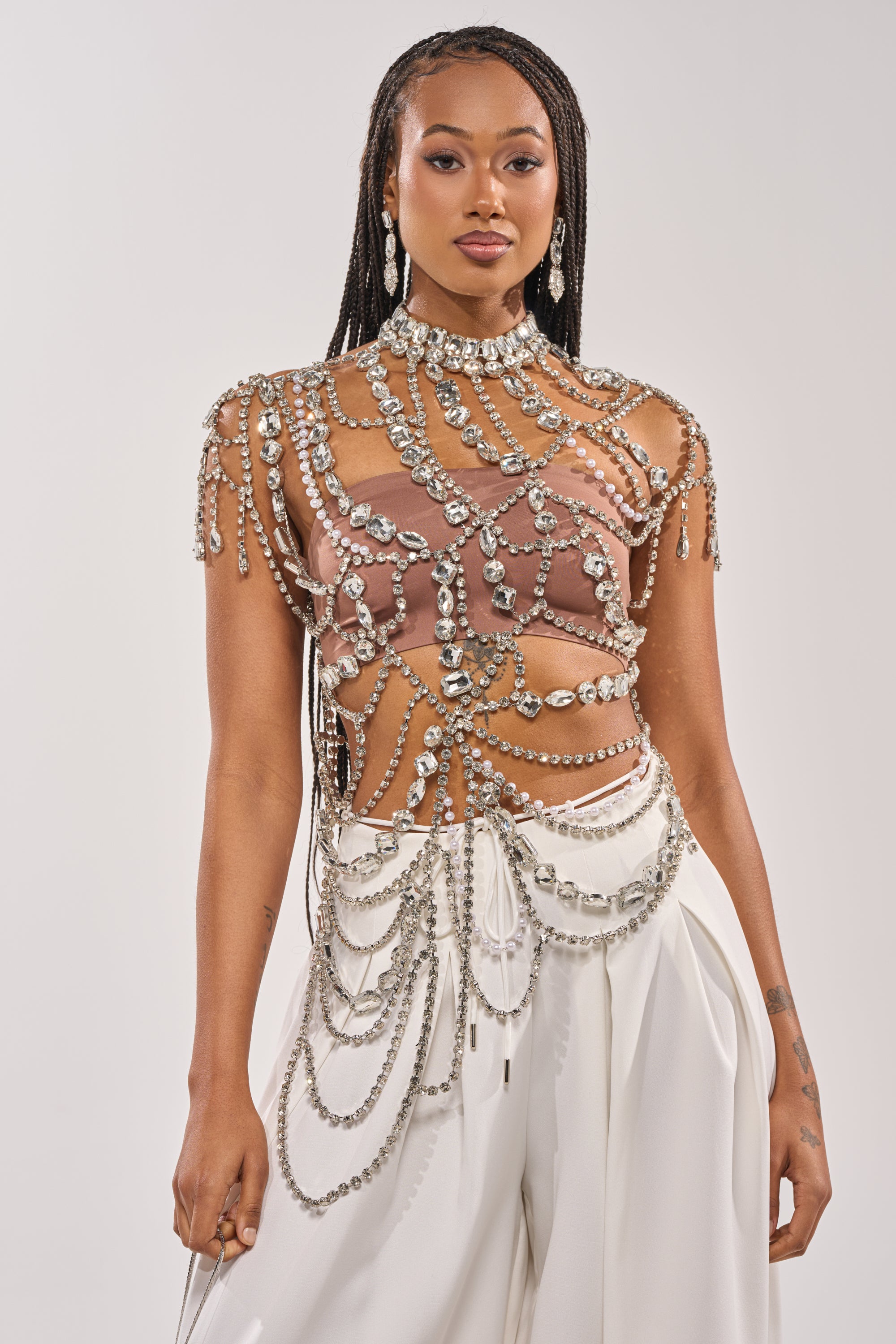 A woman with long braids models the REGAL RHINESTONE CHAIN TOP, featuring sparkling crystal chains and large gems over a mauve bralette and high-waisted white pants, striking a glamorous pose against a plain background. 0