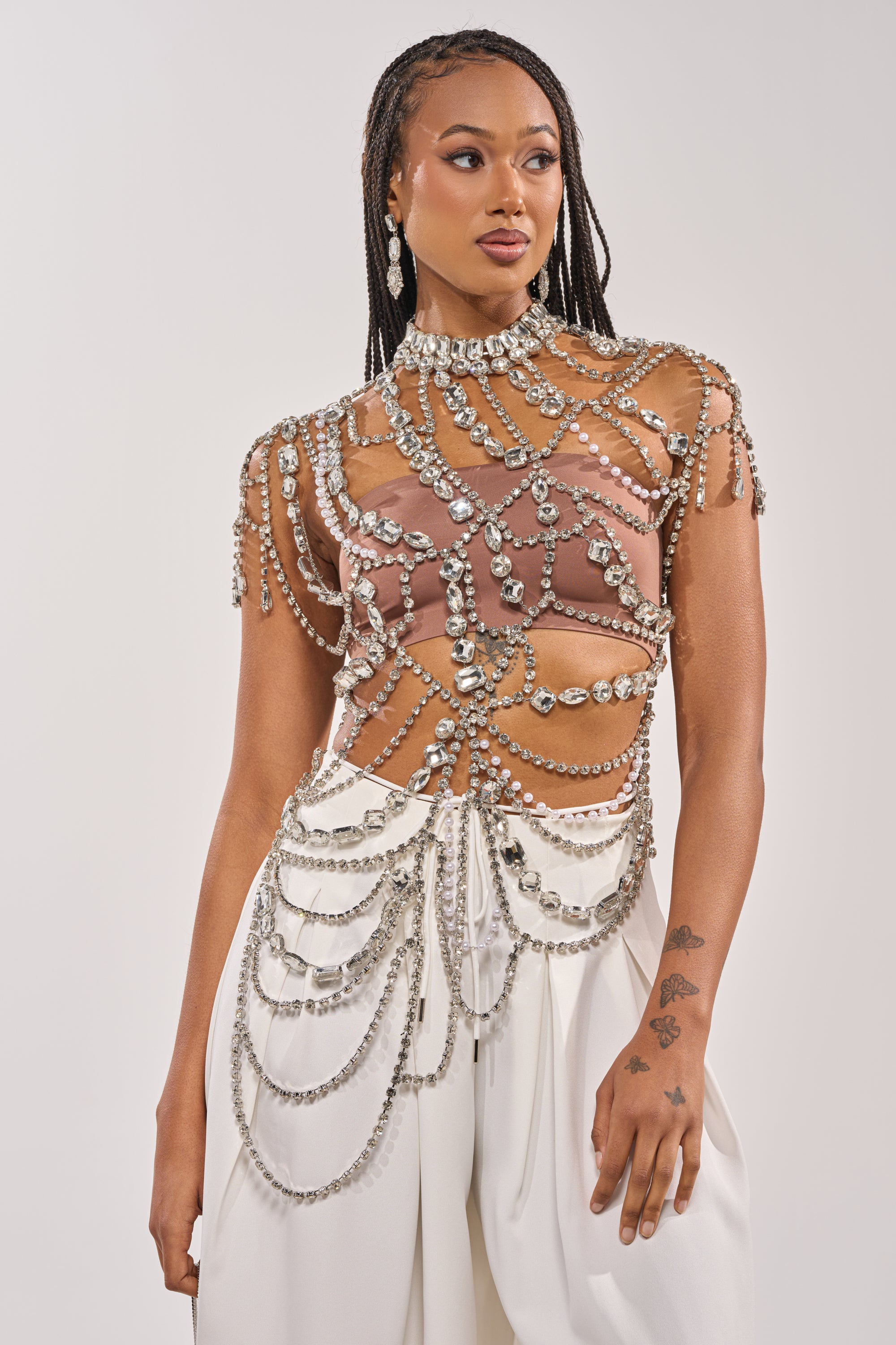 A woman with long braids wears the REGAL RHINESTONE CHAIN TOP—sheer, sparkling with crystals and pearls—paired with high-waisted white pants. She looks left, displaying dangling earrings and a hand tattoo. 1