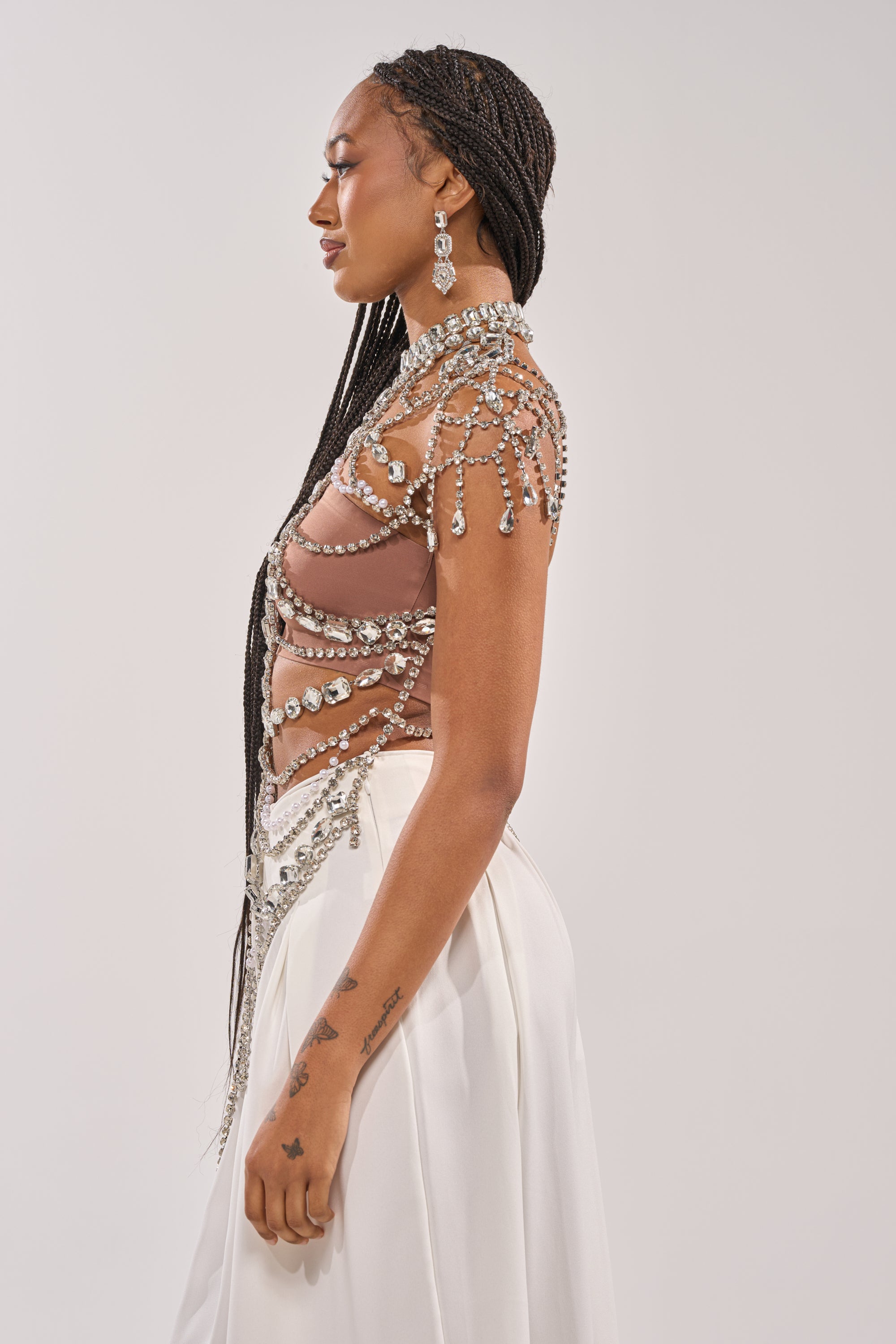 A woman with long braids and visible right arm tattoos poses in profile against a light backdrop, wearing the REGAL RHINESTONE CHAIN TOP and a white skirt. She accessorizes with dangling earrings, radiating high-glam elegance. 3