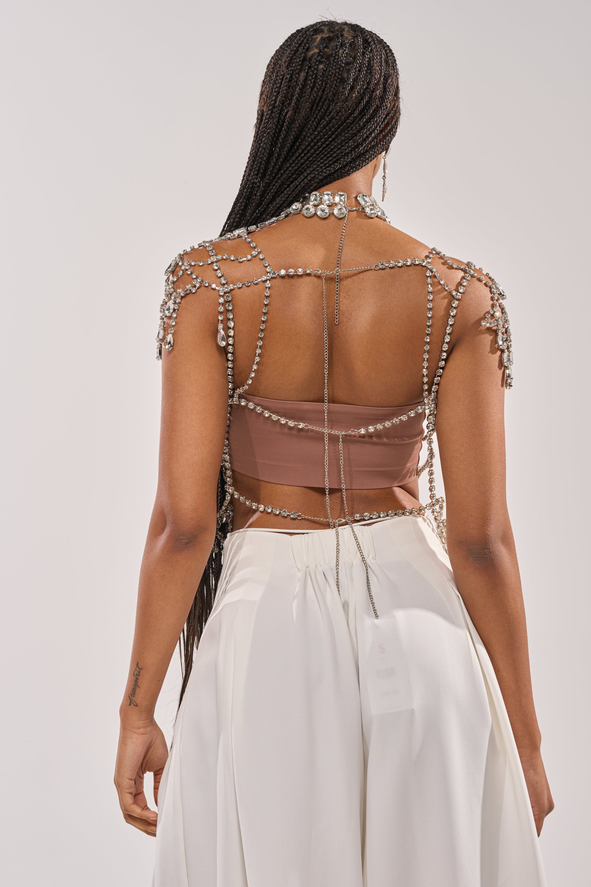 A woman with long braids is seen from behind wearing the REGAL RHINESTONE CHAIN TOP over a brown strapless top and white high-waisted pants, against a plain light background. 4