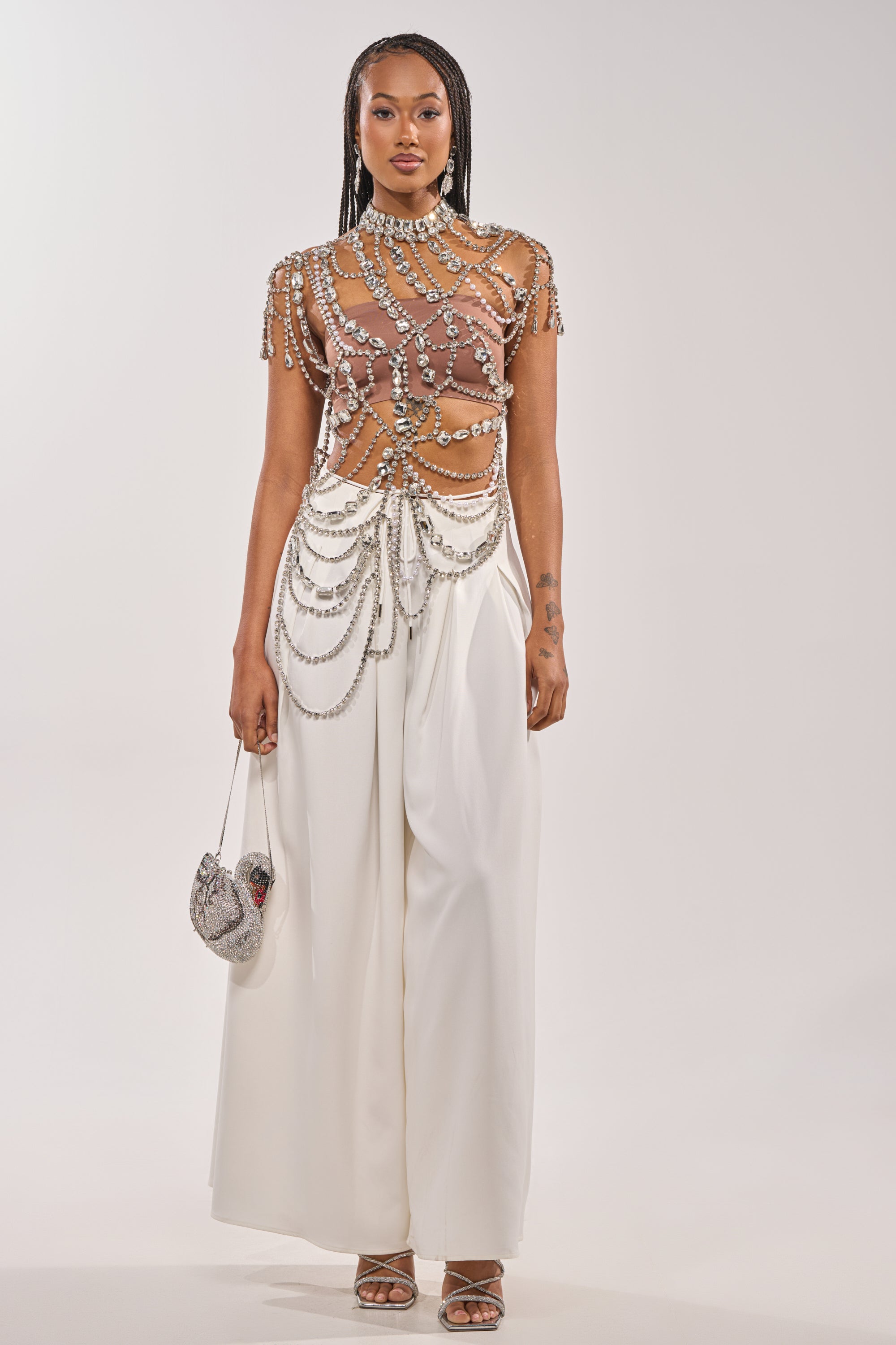A woman wears the REGAL RHINESTONE CHAIN TOP with pearl chains, wide-legged white pants, and heeled sandals. She carries a small silver beaded handbag and has long, braided hair. 5