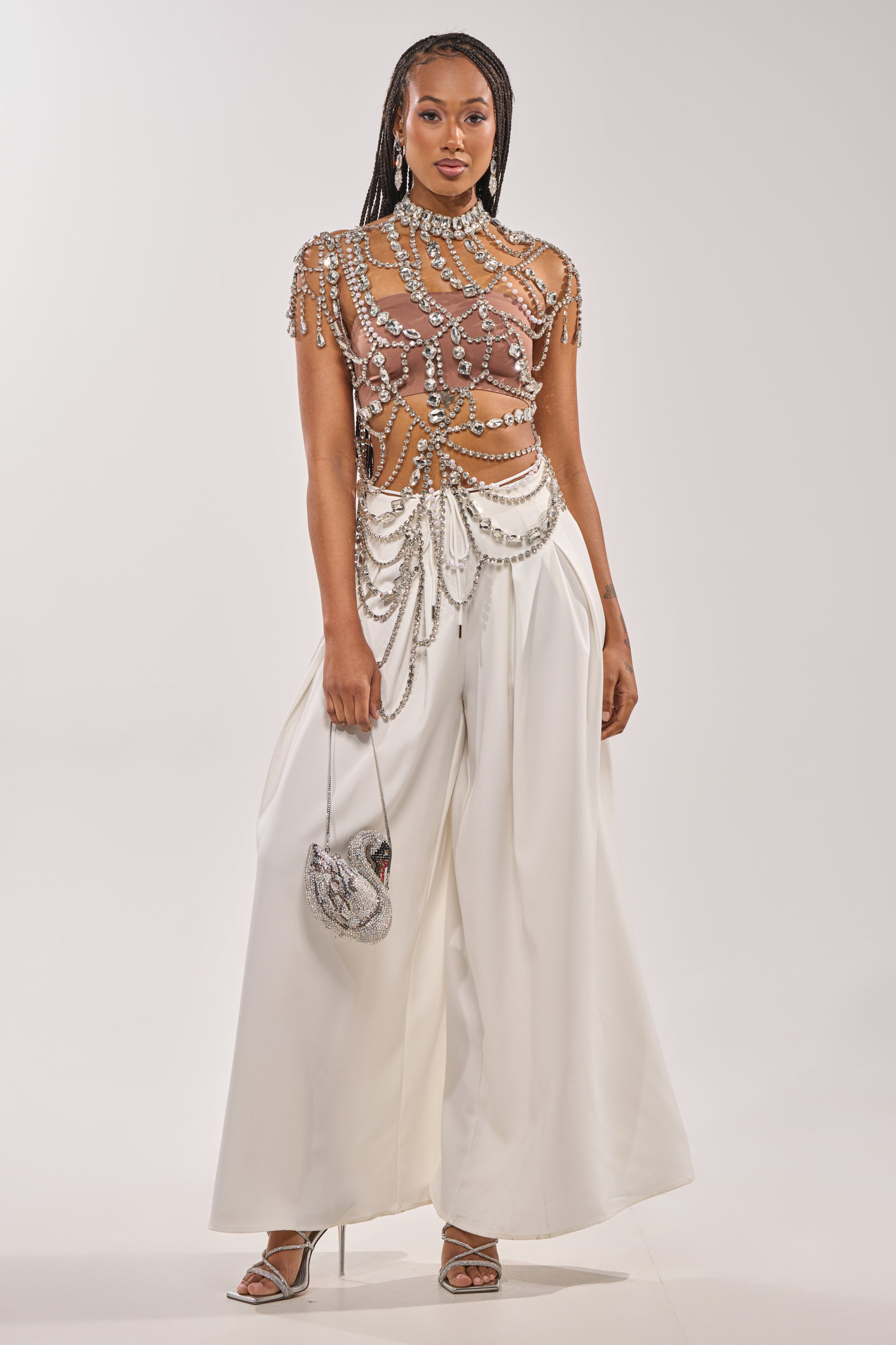 A woman stands confidently in the REGAL RHINESTONE CHAIN TOP, styled with wide-legged white pants, silver heels, and a chic silver purse against a plain light background. 6