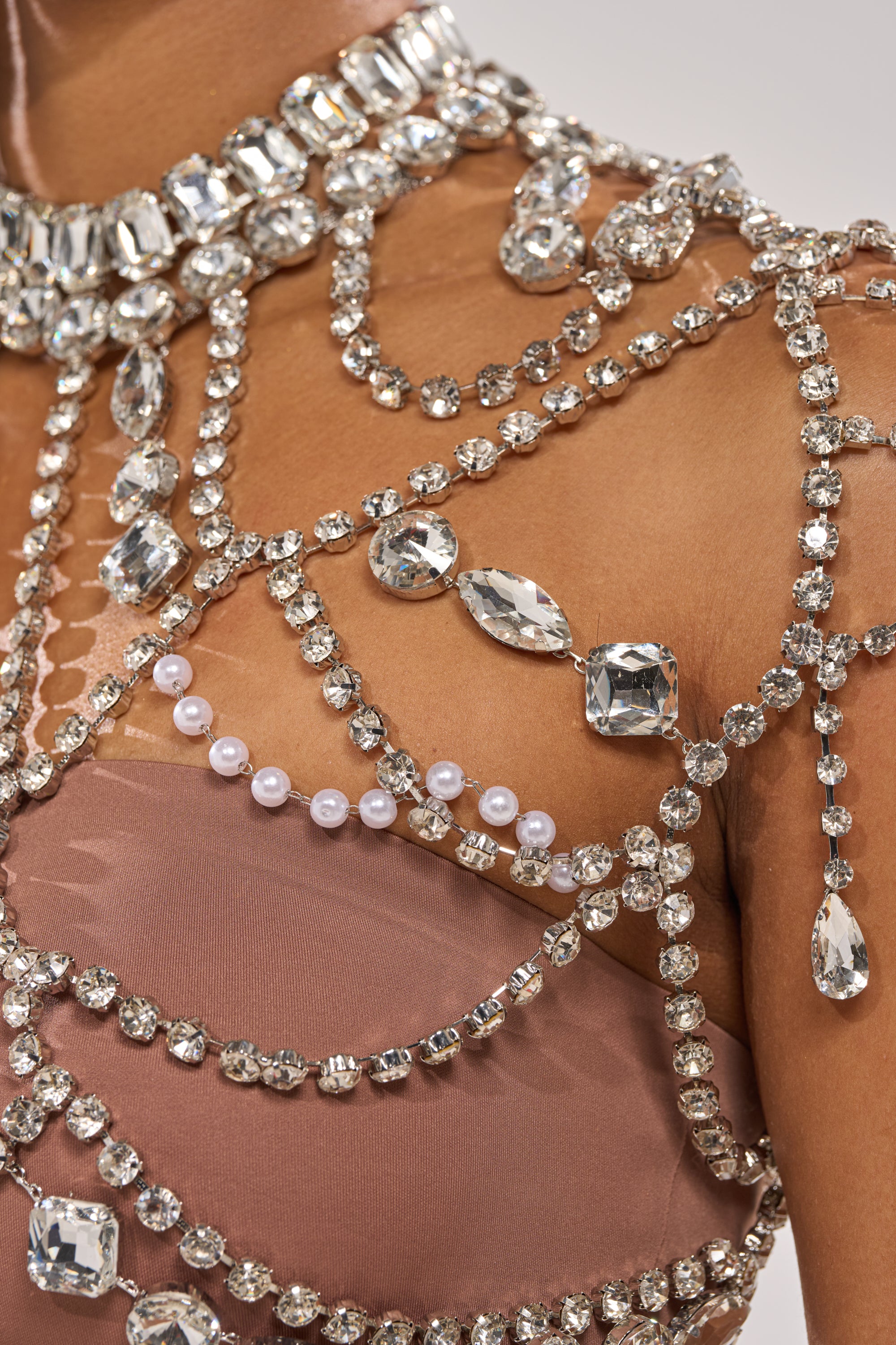 A close-up of a person wearing the REGAL RHINESTONE CHAIN TOP, featuring sheer blush fabric with draped strands of sparkling silver rhinestones and pearls cascading across the chest and shoulder. 7