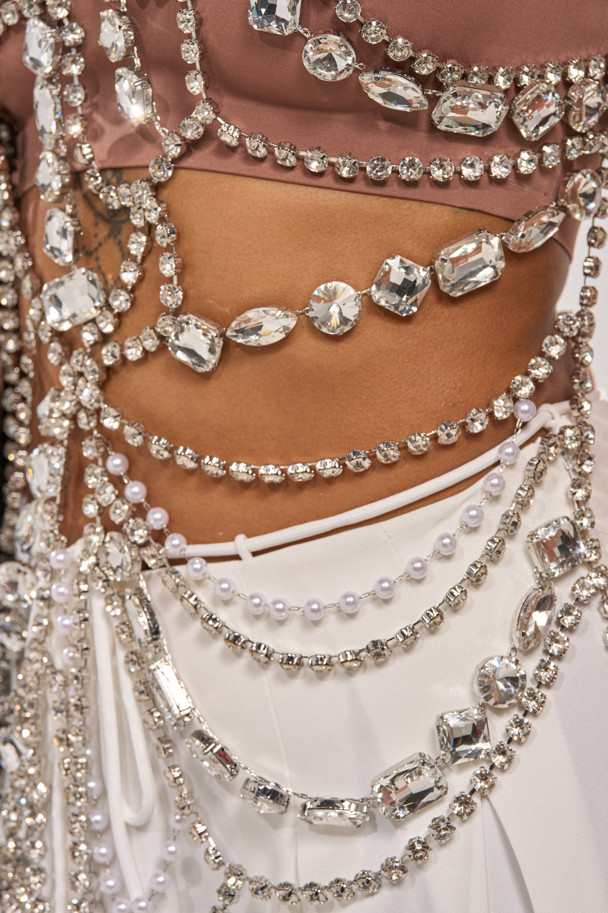 Close-up of a person wearing the REGAL RHINESTONE CHAIN TOP, featuring draped sparkling crystals, pearls, and gems over a light pink top and white fabric for an intricate, glamorous look. 8