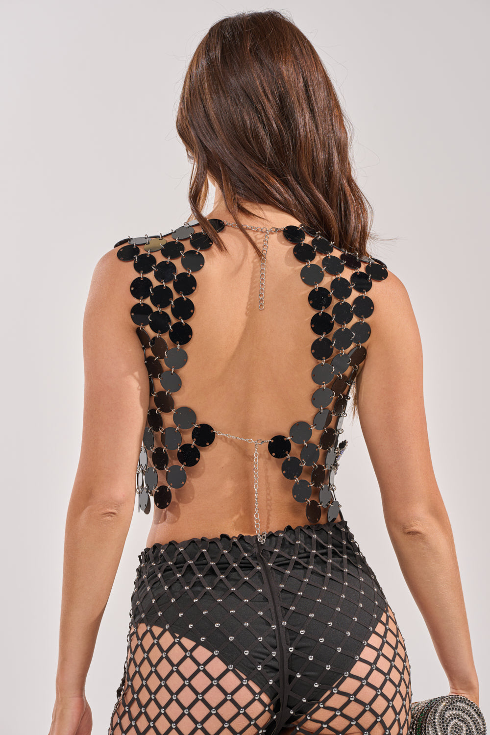 A woman with long brown hair models the KARISSA SEQUIN CHAIN TOP, featuring large black sequin discs linked by chains, paired with high-waisted fishnet bottoms. She is shown from the back against a neutral backdrop. 3