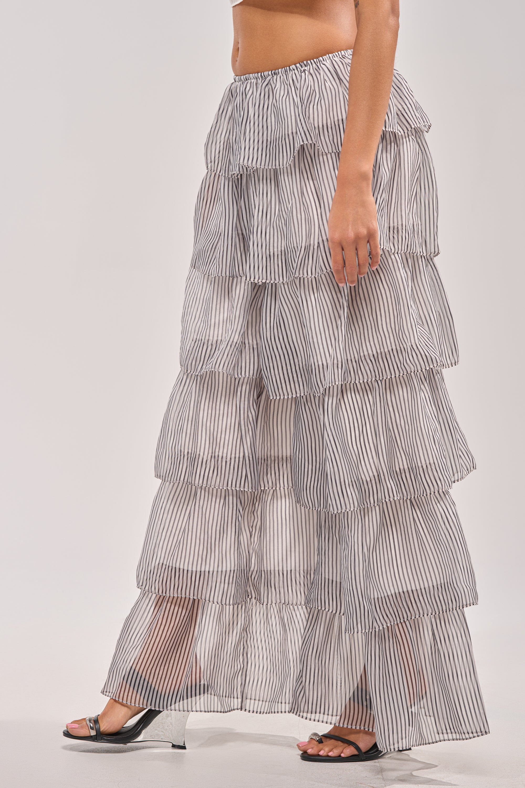 A woman wears the ALL I WANT TO DO STRIPE PALAZZO PANT—ankle-length with thin vertical black and white stripes—paired with black open-toe heeled sandals, creating an elegant spring festival look. Photo cropped from waist down. 2