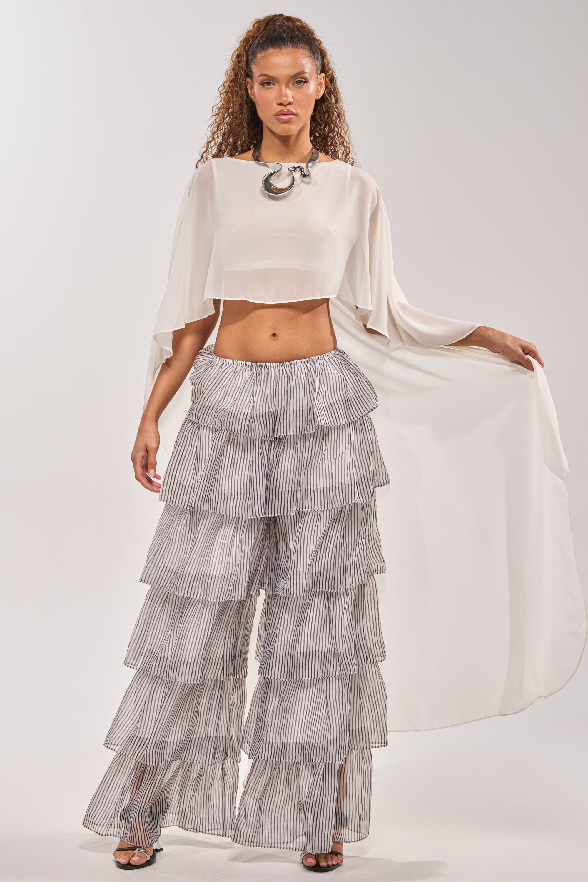 A woman models the ALL I WANT TO DO STRIPE PALAZZO PANT, styled with a white cape top, black open-toe heels, and a bold silver necklace—a striking spring festival look with layered ruffles and wide sleeves. 4