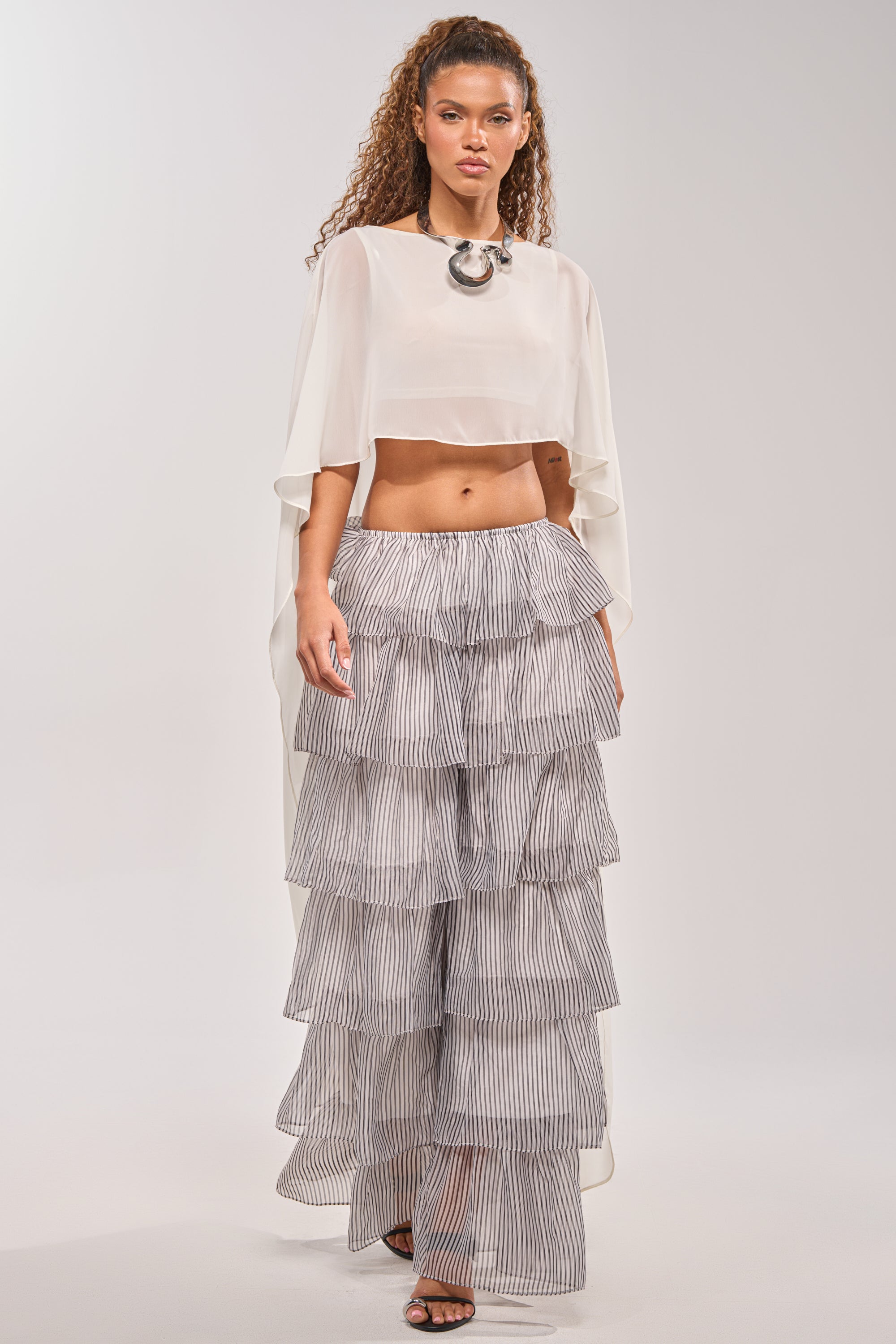 A woman models the ALL I WANT TO DO STRIPE PALAZZO PANT in gray and white with a sheer white crop top, flowy sleeves, and a statement necklace, creating the ideal spring festival look against a simple light backdrop. 5