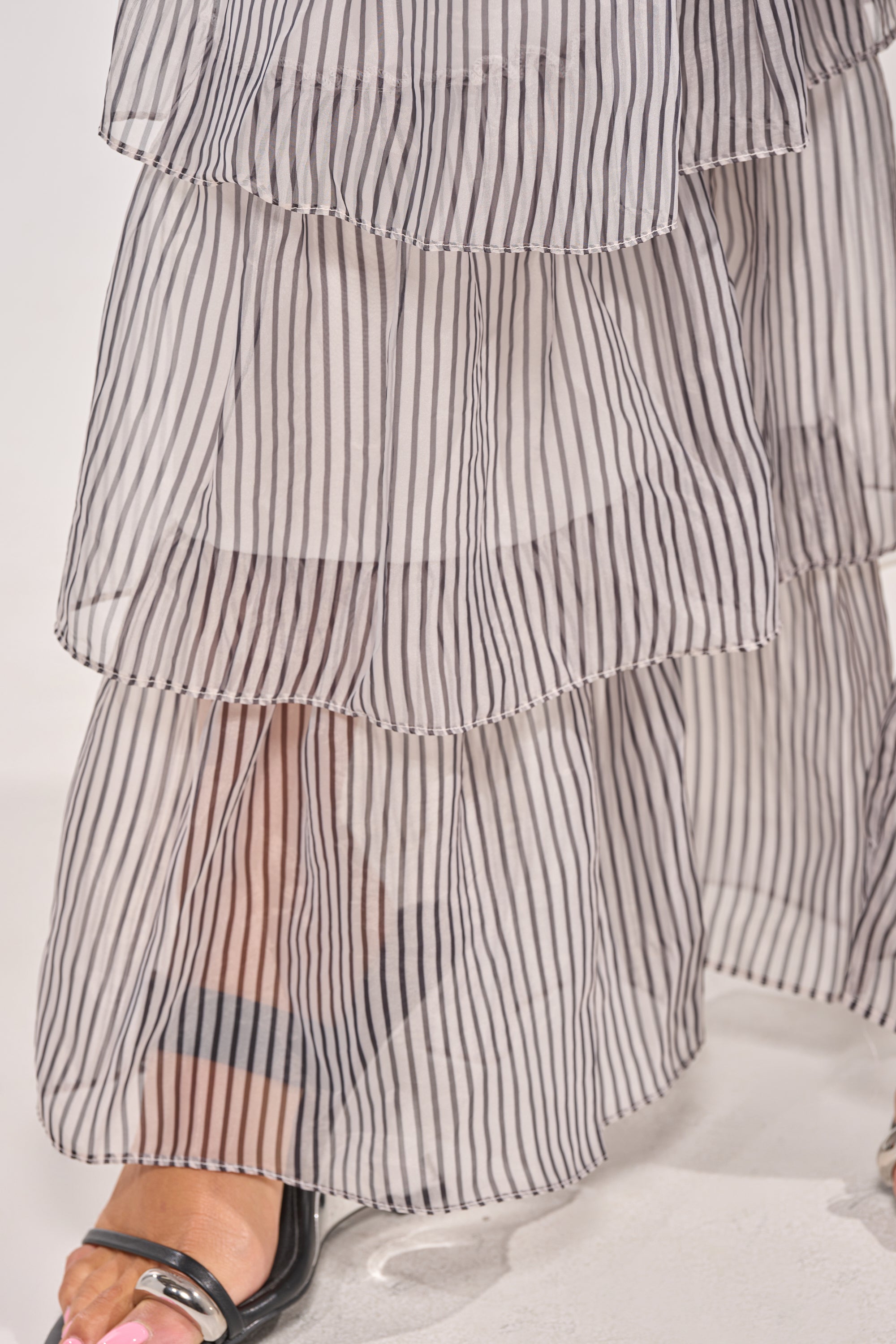 A close-up of the ALL I WANT TO DO STRIPE PALAZZO PANT, featuring sheer, layered fabric with vertical black and white stripes—perfect for spring styling—revealing the wearer’s legs and feet in black sandals. 6