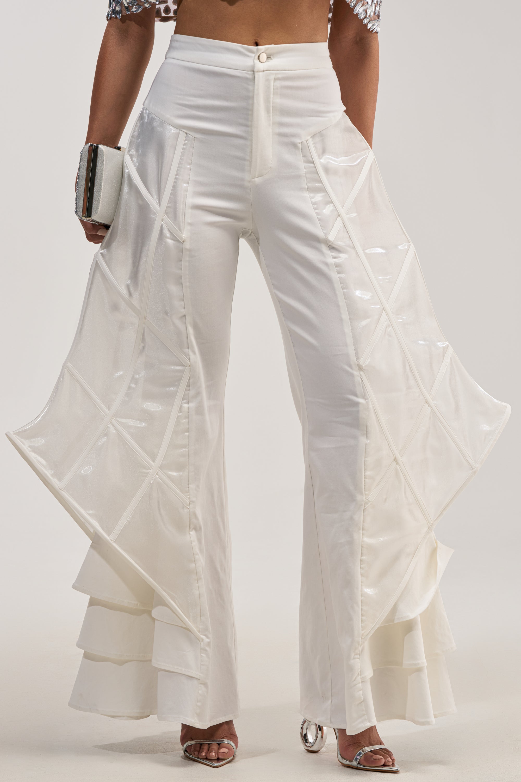 Wearing THE CRAZIER THE BETTER TROUSER, which features white flare-leg styling with sheer, flared side panels, ruffled layers at the bottom hem, and sculptural cage detailing, this person completes their look by holding a small clutch.