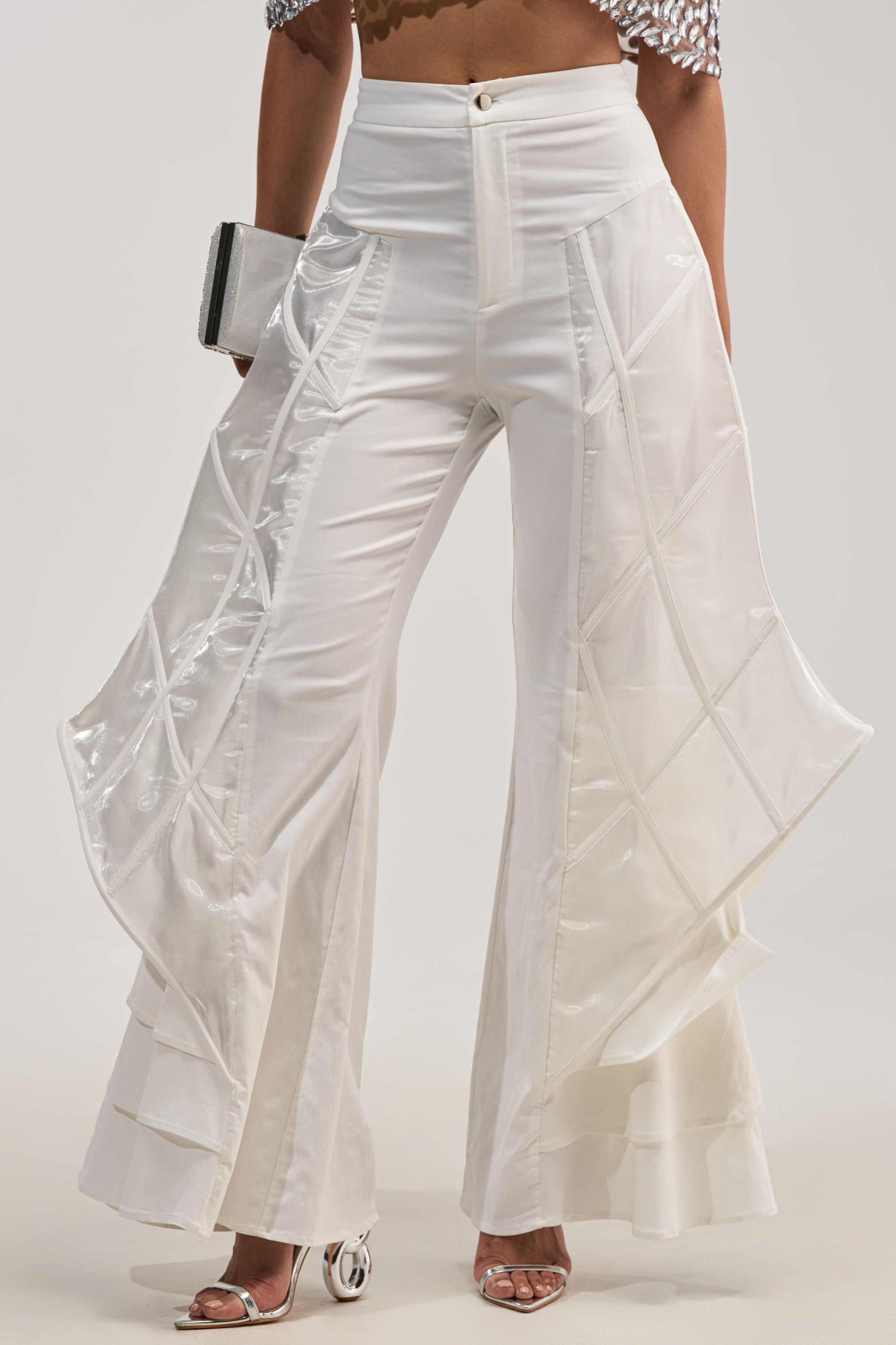 Woman wearing THE CRAZIER THE BETTER TROUSER—high-waisted, wide-legged white flare leg trousers featuring layered, flared panels with geometric patterns and delicate sculptural cage detailing—paired with silver heels and a metallic clutch.
