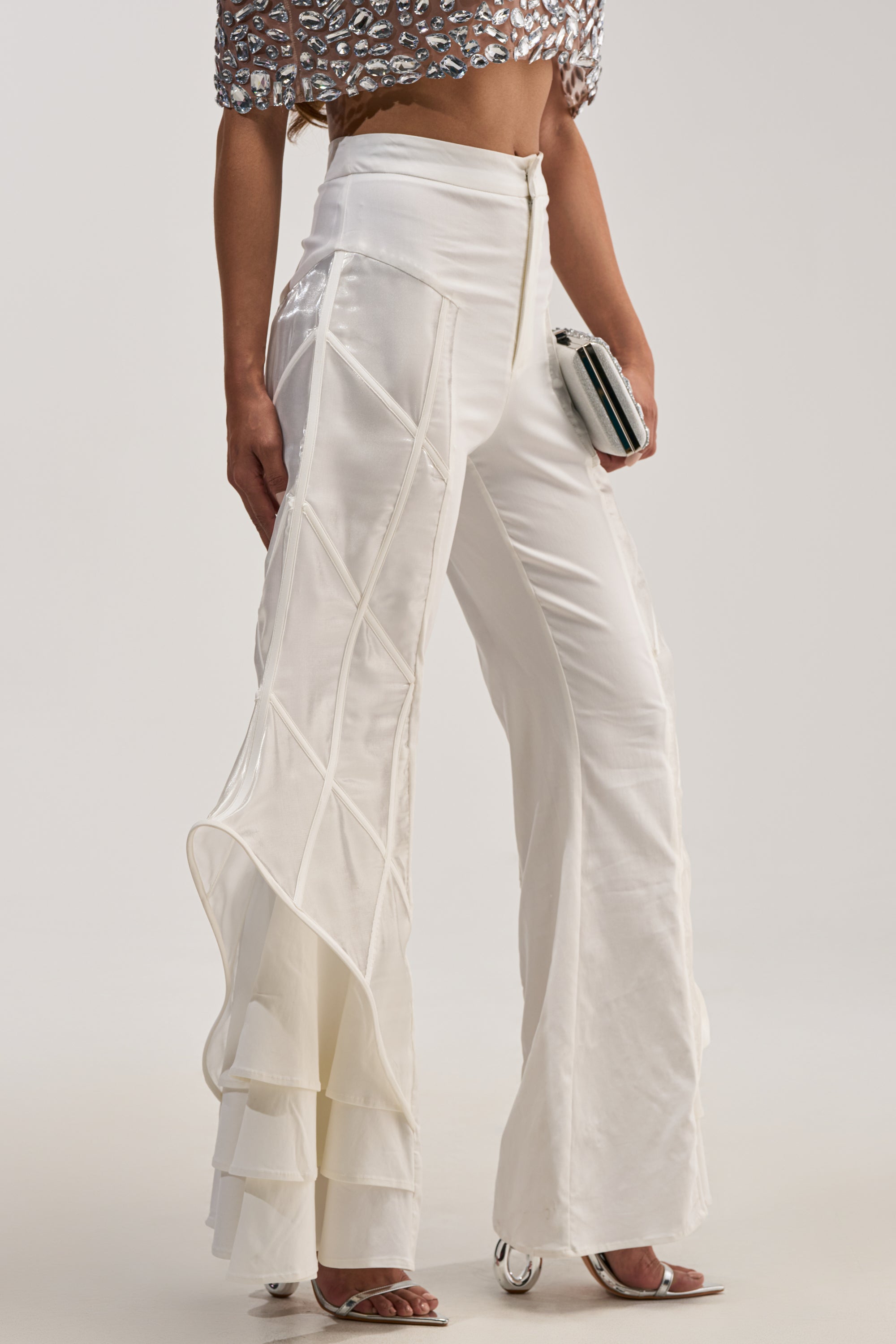 Wearing THE CRAZIER THE BETTER TROUSER—high-waisted white flare leg trousers with sheer panels and sculptural cage detailing—and a shiny crop top, a person holds a small clutch, with only their lower body visible.