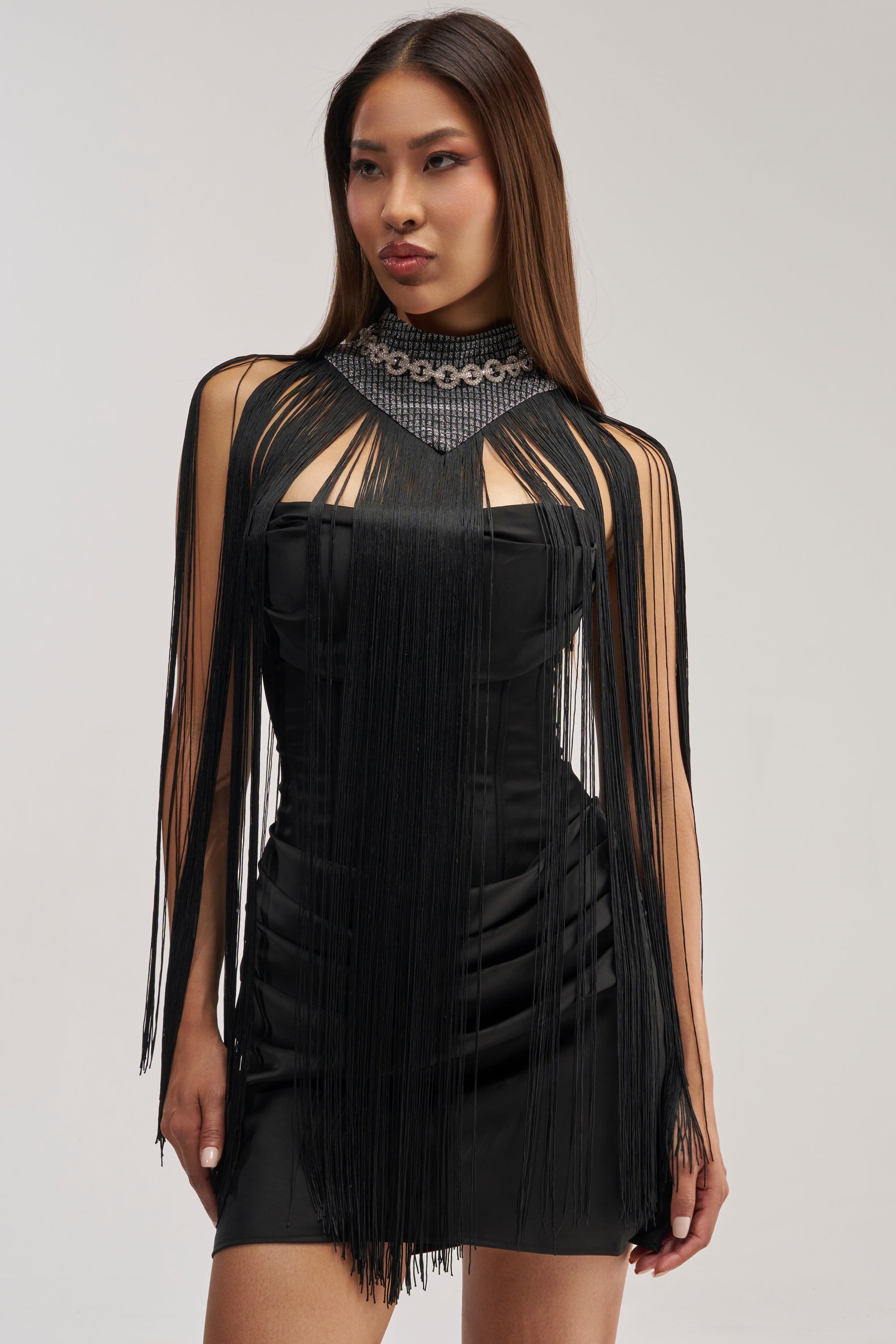 A woman stands against a plain light background, wearing a fitted black dress with long black fringe details and a geometric neckline. Her straight hair frames her elegant look, which she completes with the I AM DRAMA NECKLACE—a rhinestone choker that adds a touch of vintage-inspired glamour to the ensemble.