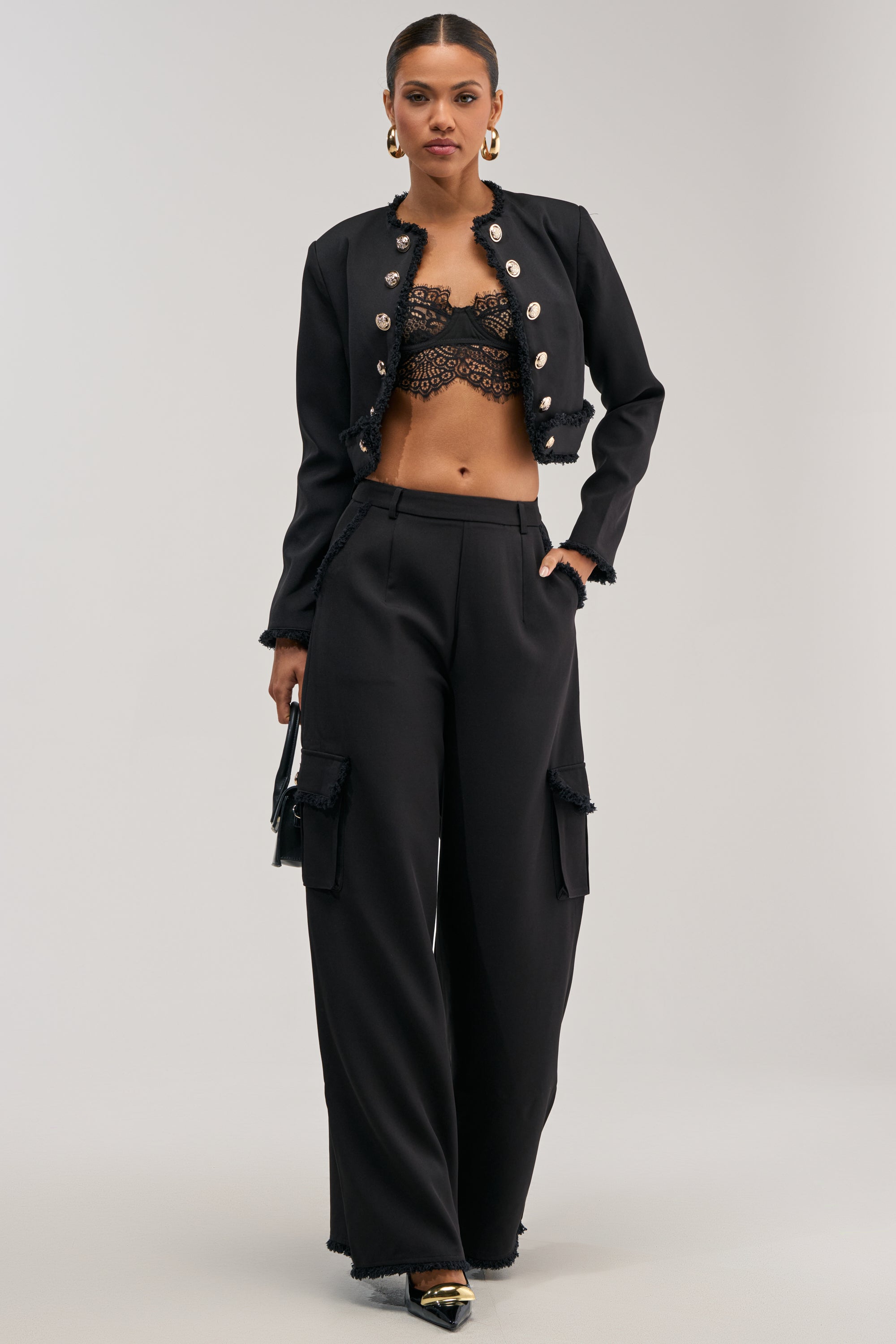 A woman stands confidently in CELINE CARGO TROUSERs, paired with a black cropped jacket with gold buttons, a black lace bralette, black heels, a small black handbag, and gold hoop earrings.