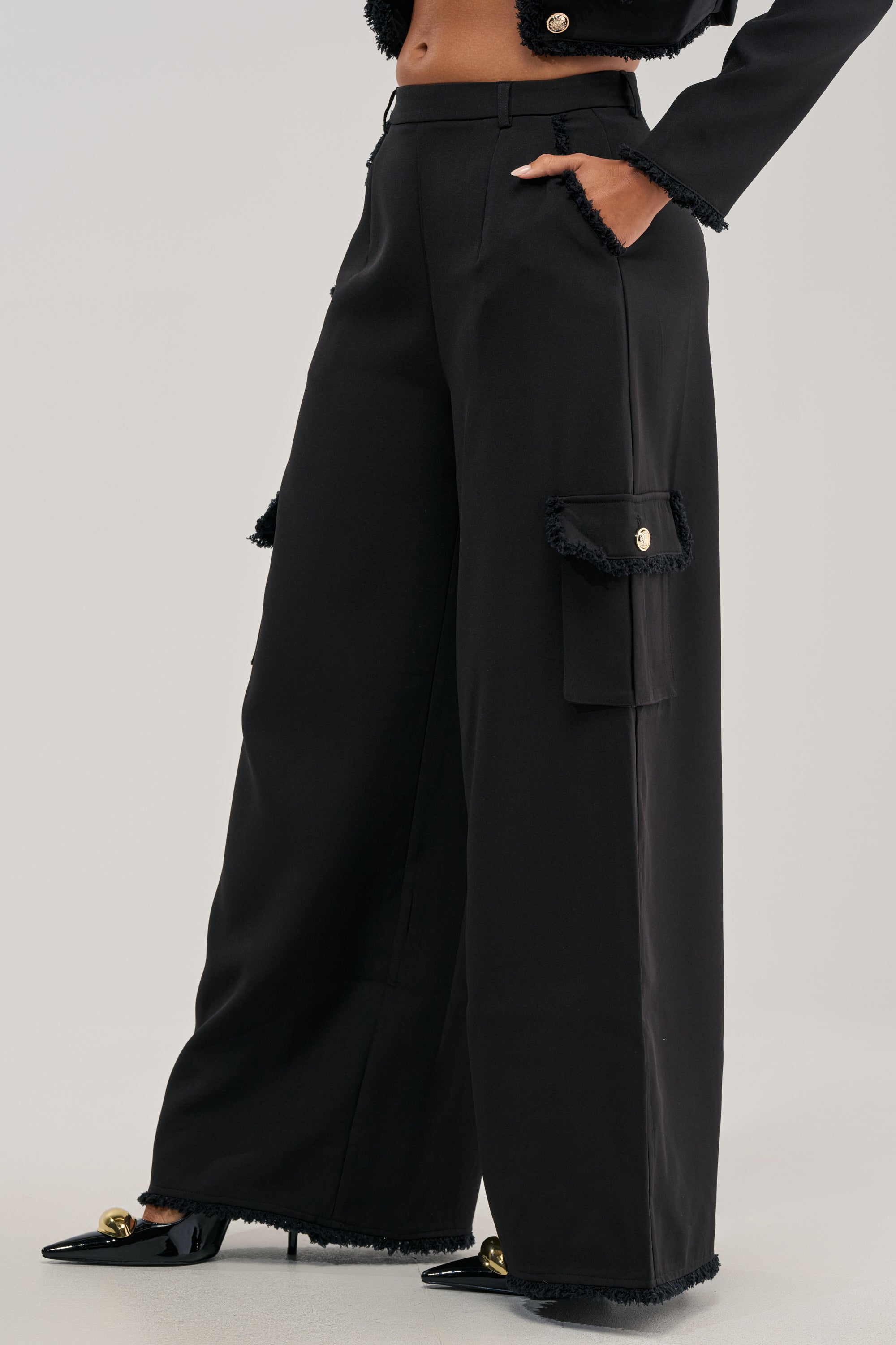 Dressed in the CLASSY VIBE CARGO TROUSER—featuring a striking high-waisted, wide-leg silhouette with black fabric and bold gold buttons—paired elegantly with black pointed-toe heels and a coordinating jacket for contemporary office sophistication.