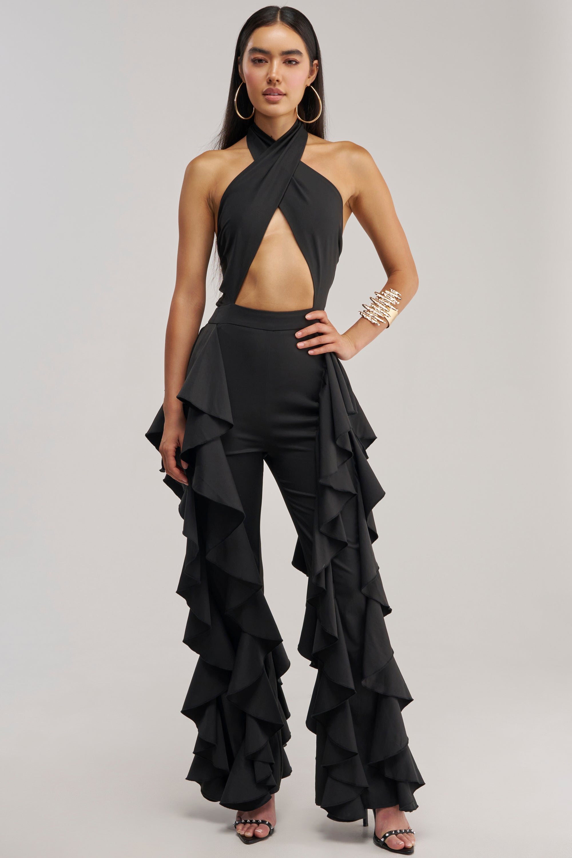 A model with long dark hair wears the DANCE BREAK RUFFLE JUMPSUIT IN BLACK—a sexy halter jumpsuit featuring a midriff cutout and dramatic ruffles cascading down the outer legs. She accessorizes with large gold hoop earrings and a wide stacked gold cuff bracelet, posing against a light gray backdrop.