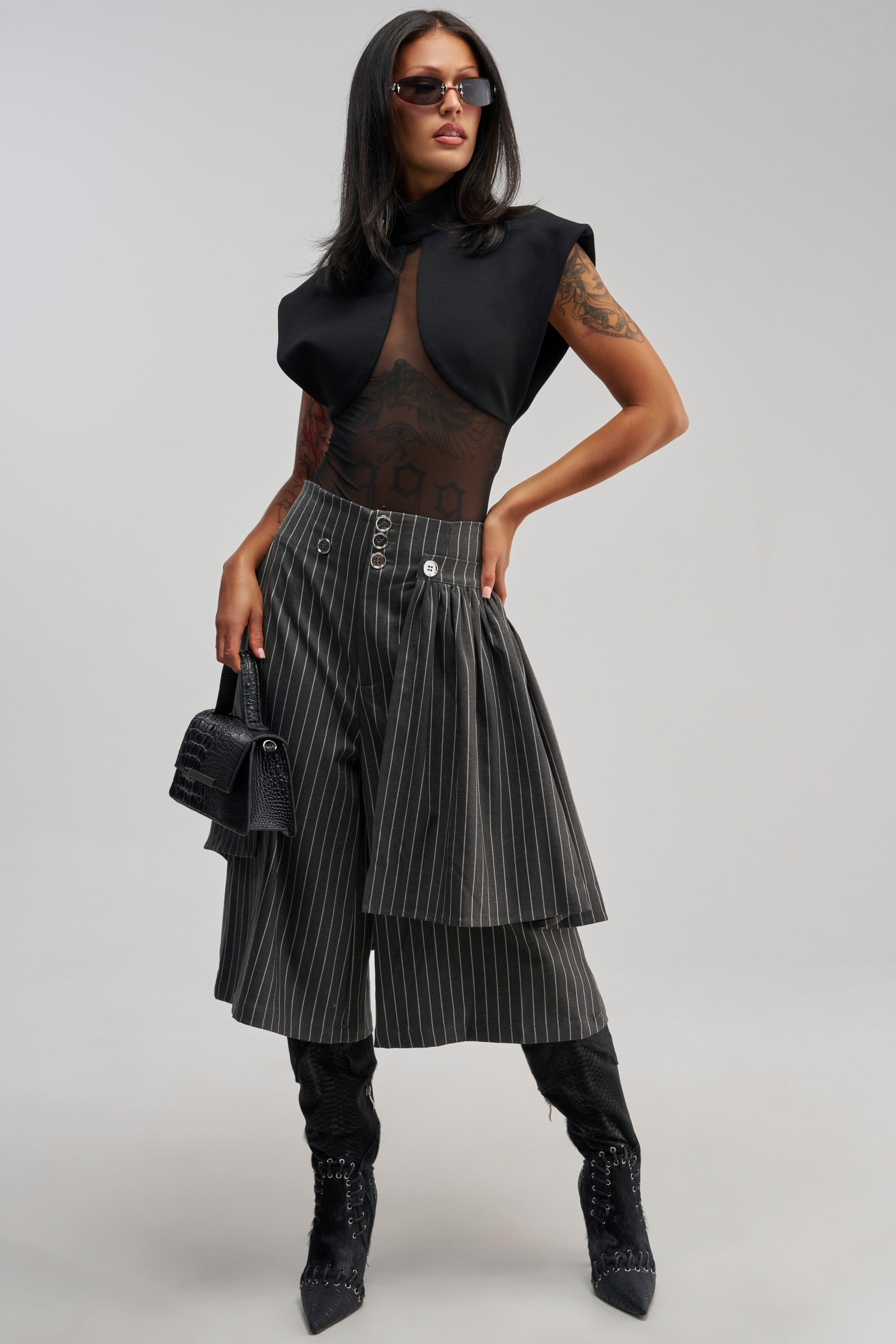 Wearing sunglasses, a woman confidently models the OFFICE SIREN STRIPED GAUCHO PANT—grey pinstripe pants with wide, cropped gaucho legs—styled with a black sheer top, black mini handbag, and black boots against a plain background for an elevated workwear look.