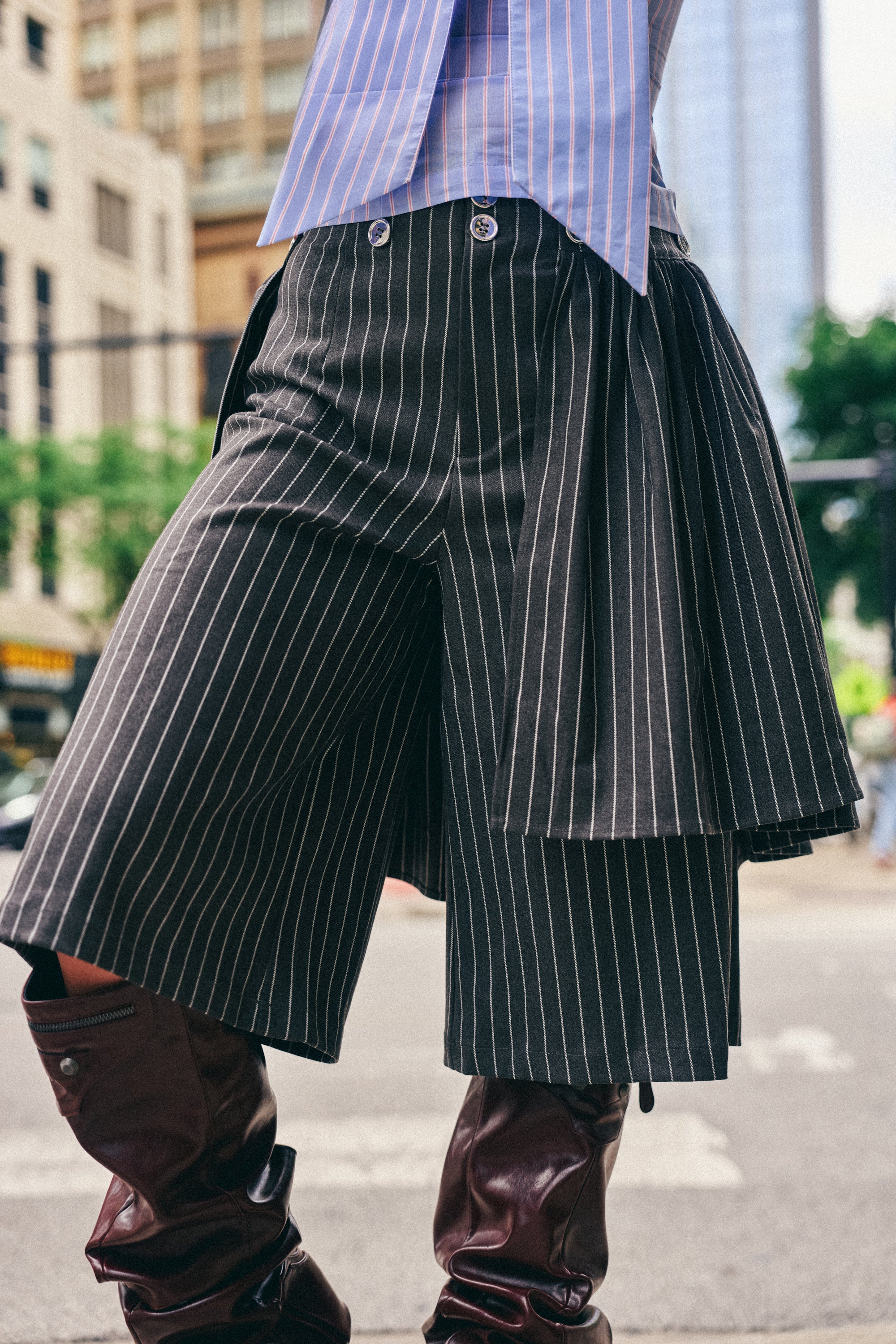 Wearing the OFFICE SIREN STRIPED GUACHO PANT, featuring a removable pleated skirt, paired with brown knee-high boots and a blue striped shirt, this individual stands on a city street, epitomizing elevated workwear.