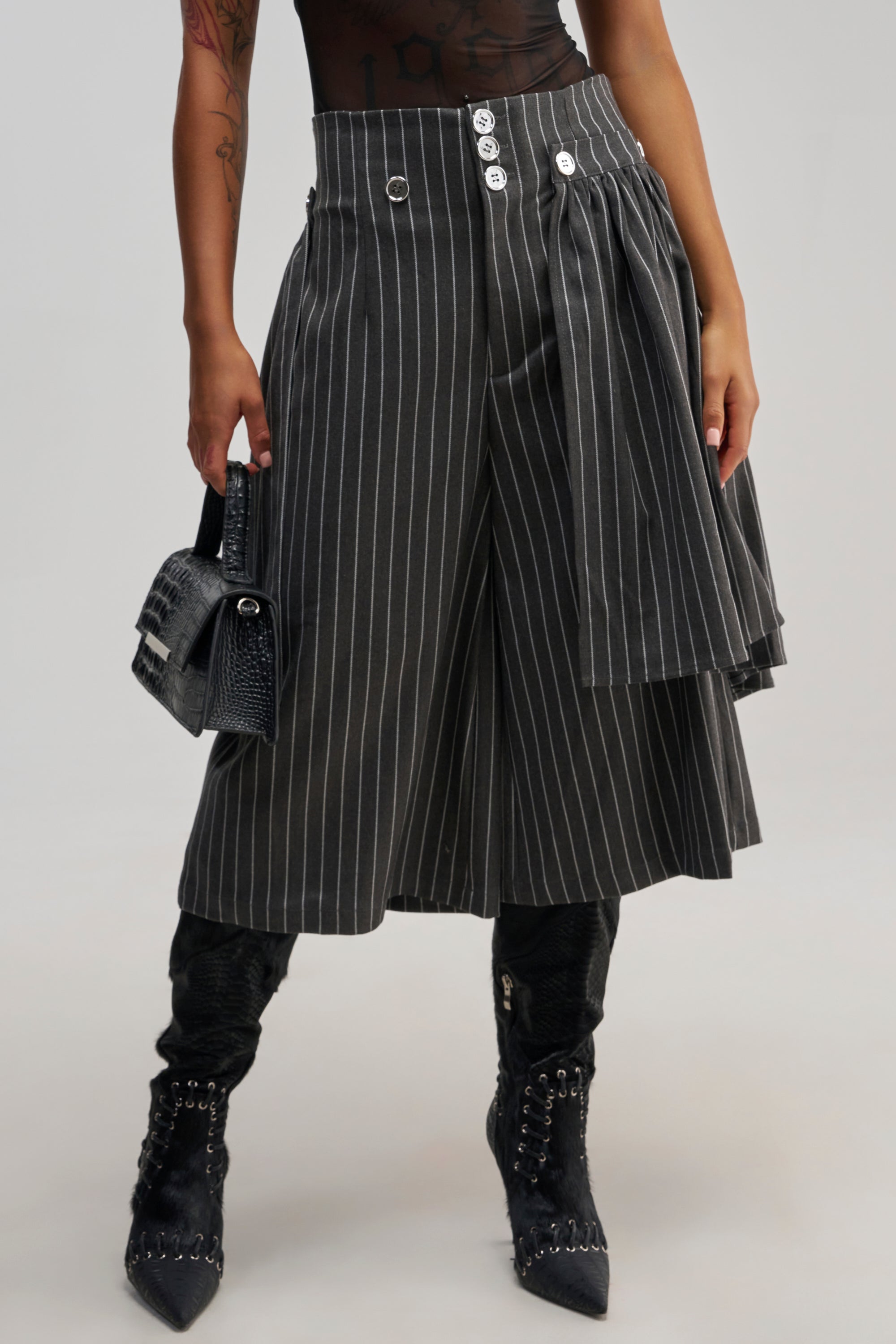 A person wears the OFFICE SIREN STRIPED GAUCHO PANT, featuring wide-leg gray pinstripes and decorative buttons, paired with black lace-up boots and a textured black handbag. The elevated workwear look is stylish and modern, highlighted by the sharp tailoring and cropped silhouette of the OFFICE SIREN STRIPED GAUCHO PANT. The background is plain and light.