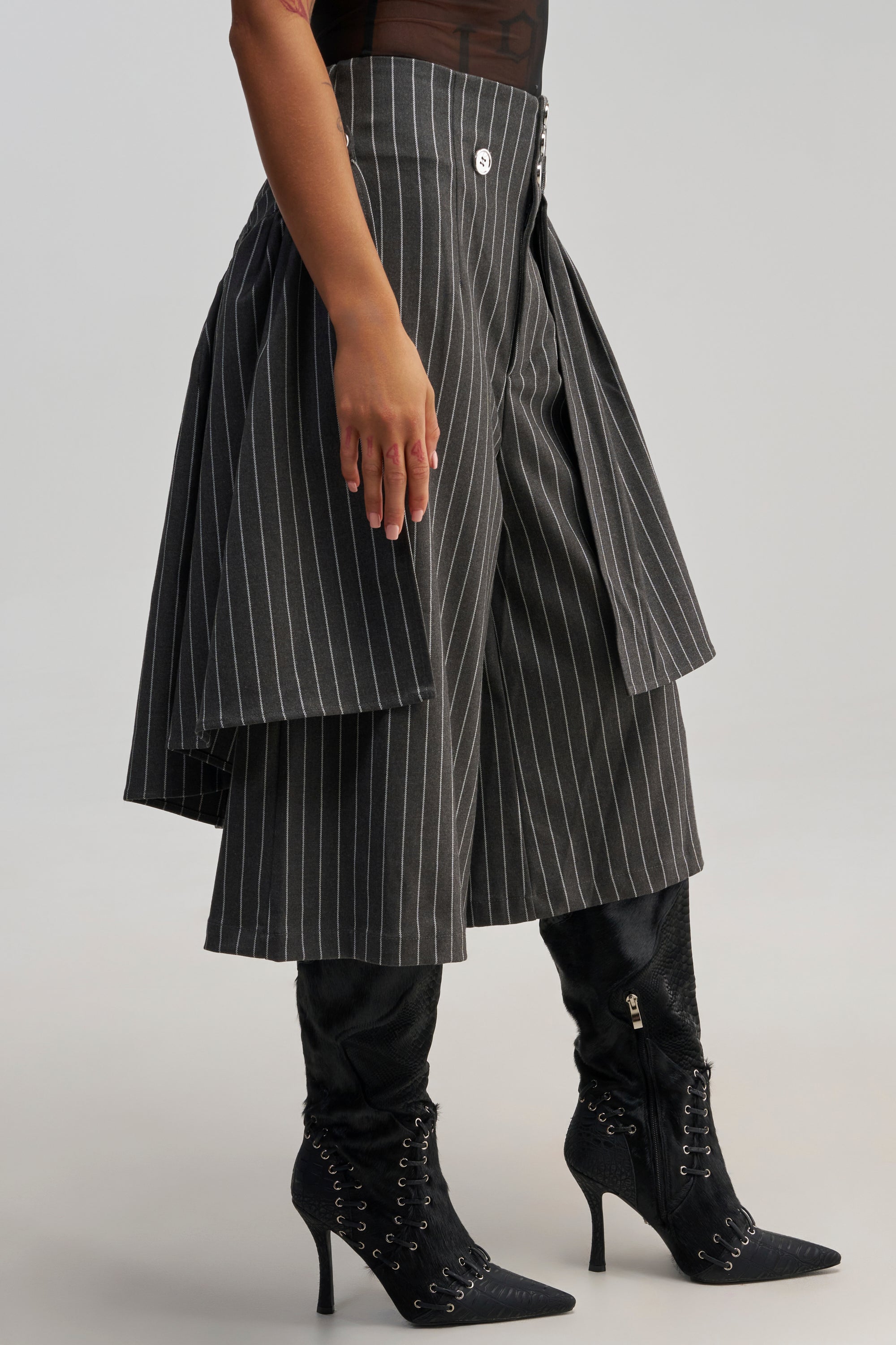 Wearing the OFFICE SIREN STRIPED GAUCHO PANT with wide, cropped pinstripe legs and layered fabric panels, styled with black heeled lace-up boots and a sheer black top for an elevated workwear look.