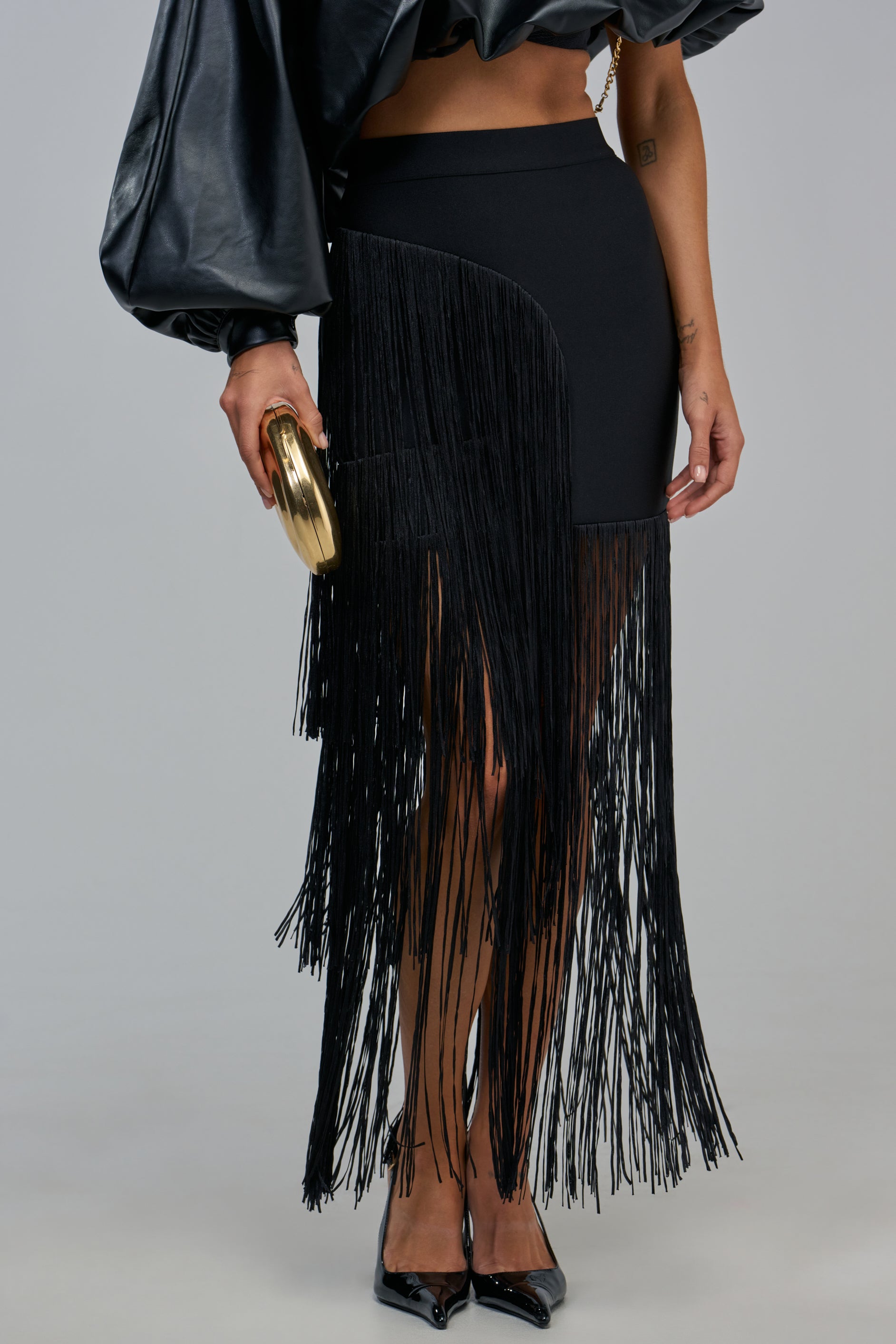 A person wears a FRINGE FOR DAYS MAXI SKIRT IN BLACK featuring long, hanging fringe on one side, paired with a black long-sleeved top and a round gold clutch.