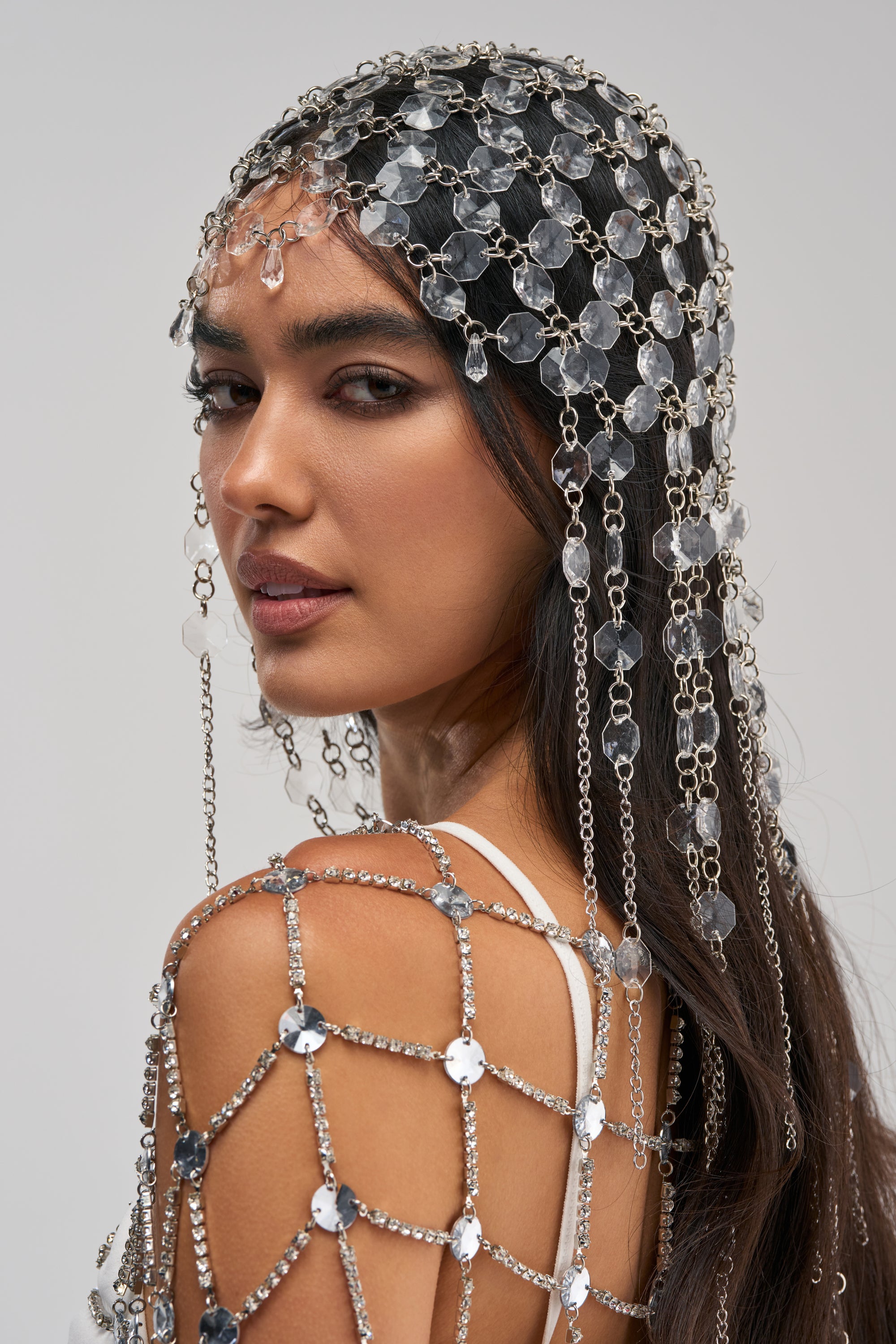 A woman with long dark hair wears the LOLLA HEAD PIECE, a striking silver chain headpiece adorned with clear crystals. She pairs it with a matching jeweled shoulder accessory, making both pieces perfect festival accessories, as she poses against a light gray background.