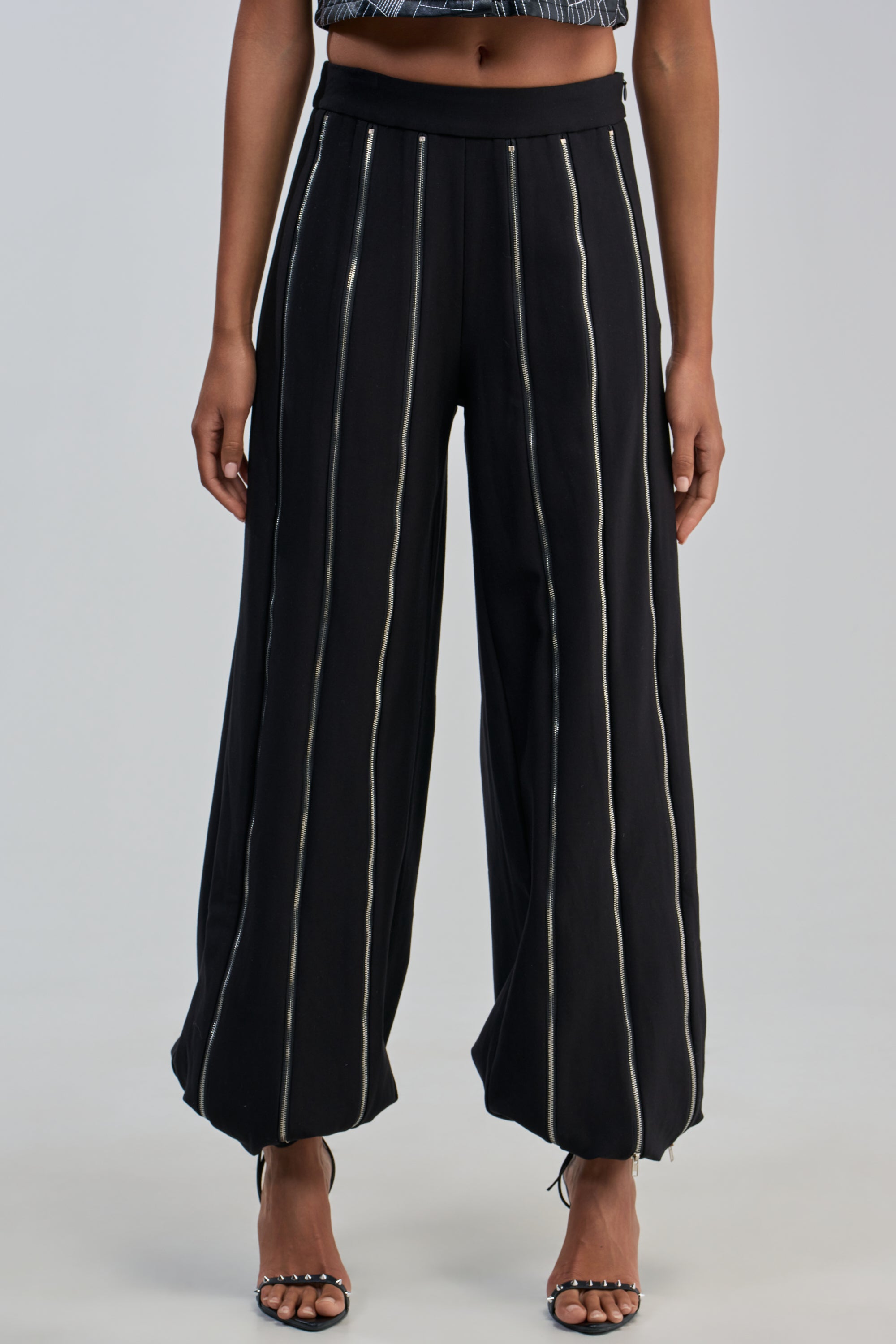 A person wearing the UNZIP ME TROUSER—black zipper balloon trousers with vertical white stripes—paired with black high-heeled sandals, standing against a plain background for a statement streetwear look that’s perfect for a rocker-chic night out.