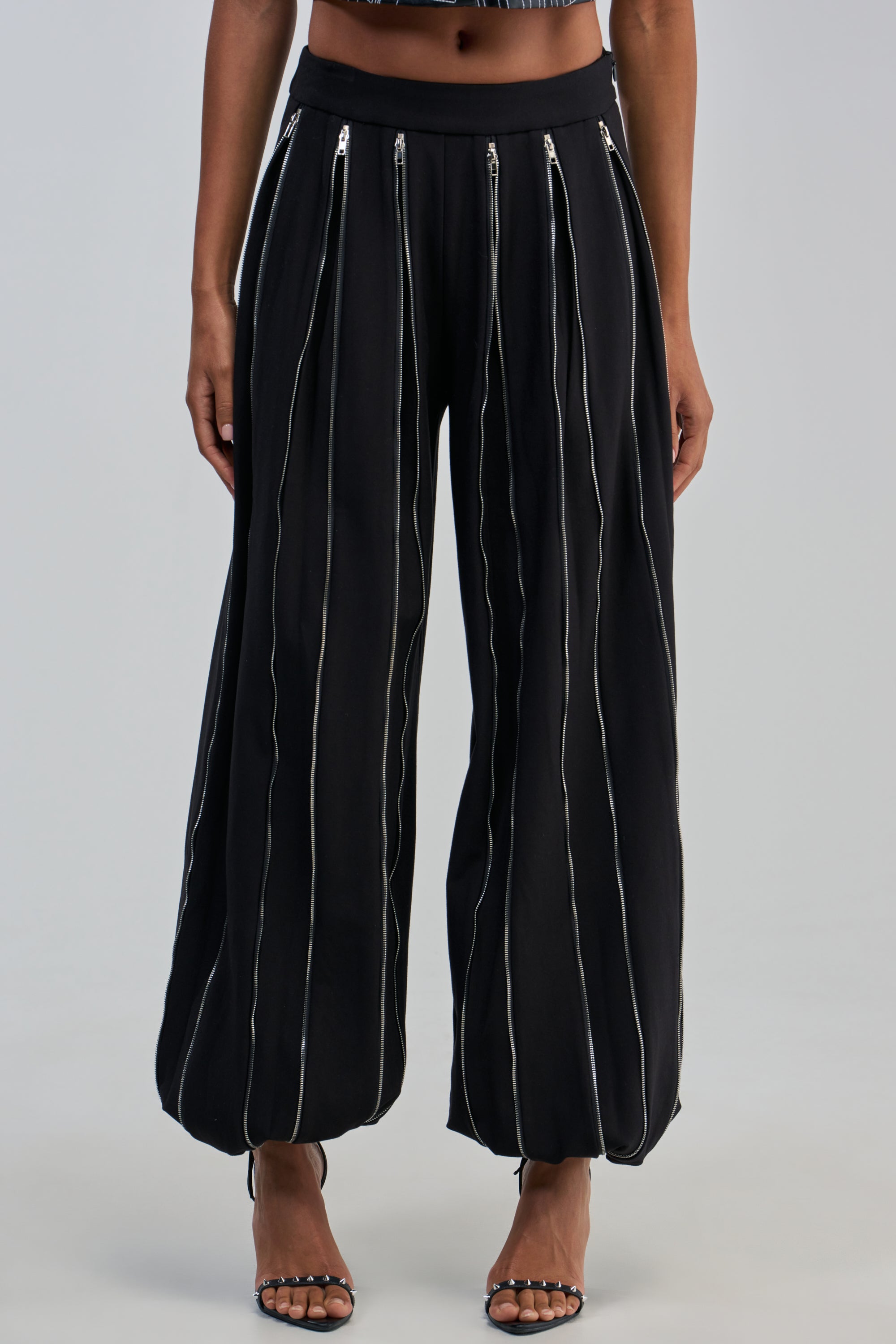 A person wearing the UNZIP ME TROUSER—high-waisted, loose-fitting black balloon pants featuring multiple vertical silver zippers down each leg—paired with studded heeled sandals for a rocker-chic night out.