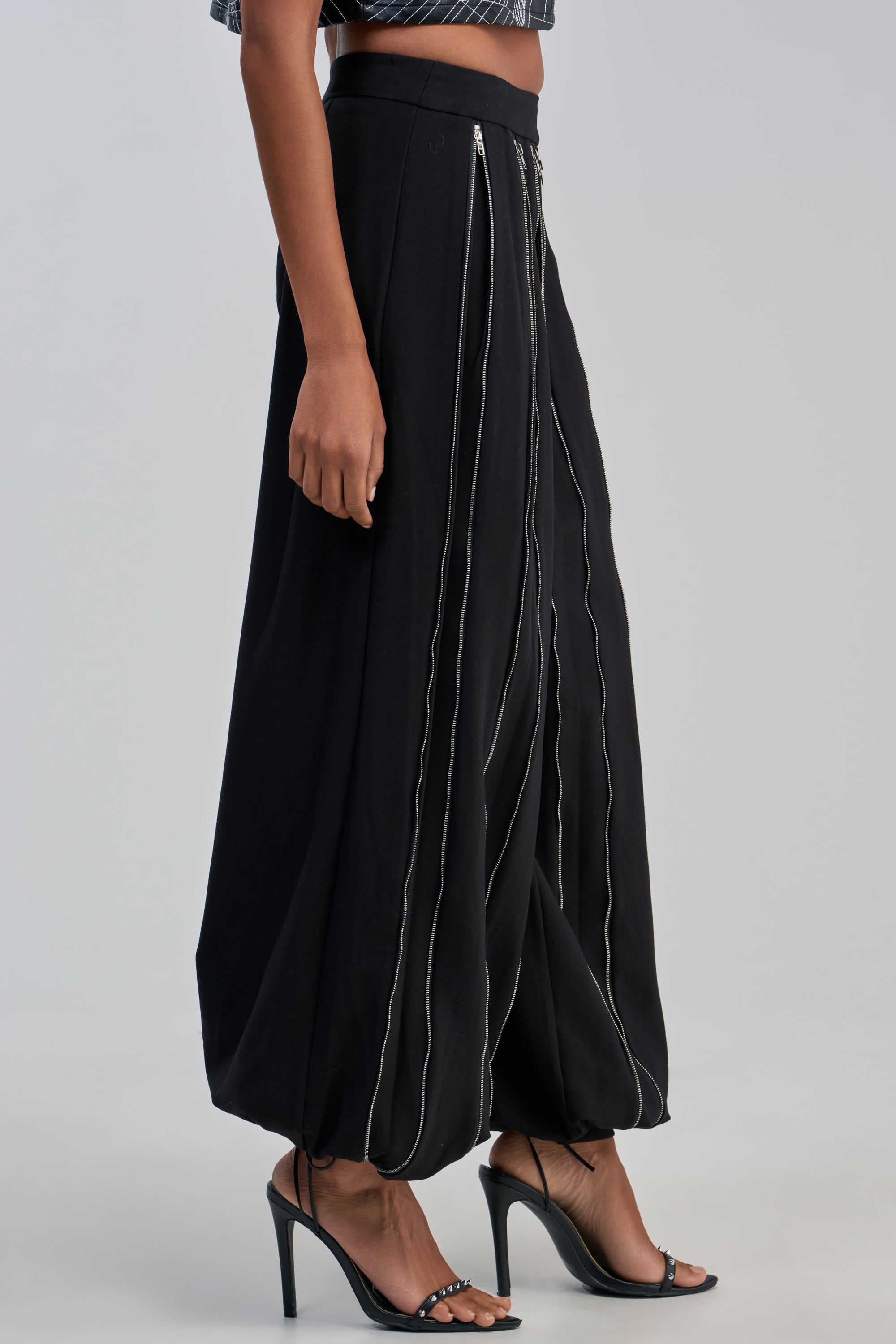 A person wearing the UNZIP ME TROUSER—loose black zipper balloon pants with white vertical stitching—paired with black high-heeled sandals, shown from the side against a plain background. This statement streetwear look is perfect for a rocker-chic night out.