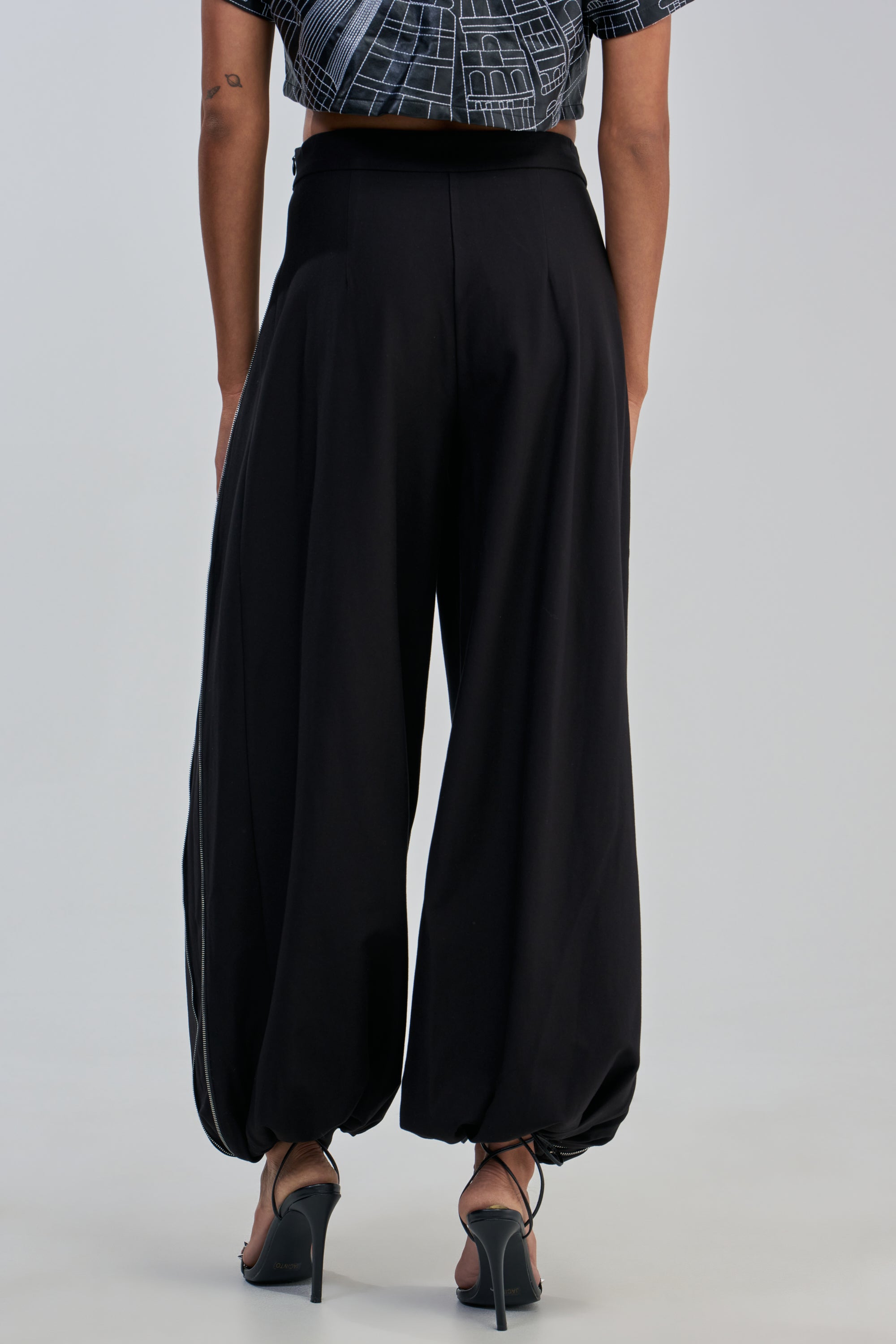 A person stands facing away, wearing the UNZIP ME TROUSER—loose-fitting black zippered balloon pants with elastic ankles—paired with black high-heeled sandals and a glimpse of a geometric-patterned black top, creating an effortlessly bold look perfect for a rocker-chic night out.