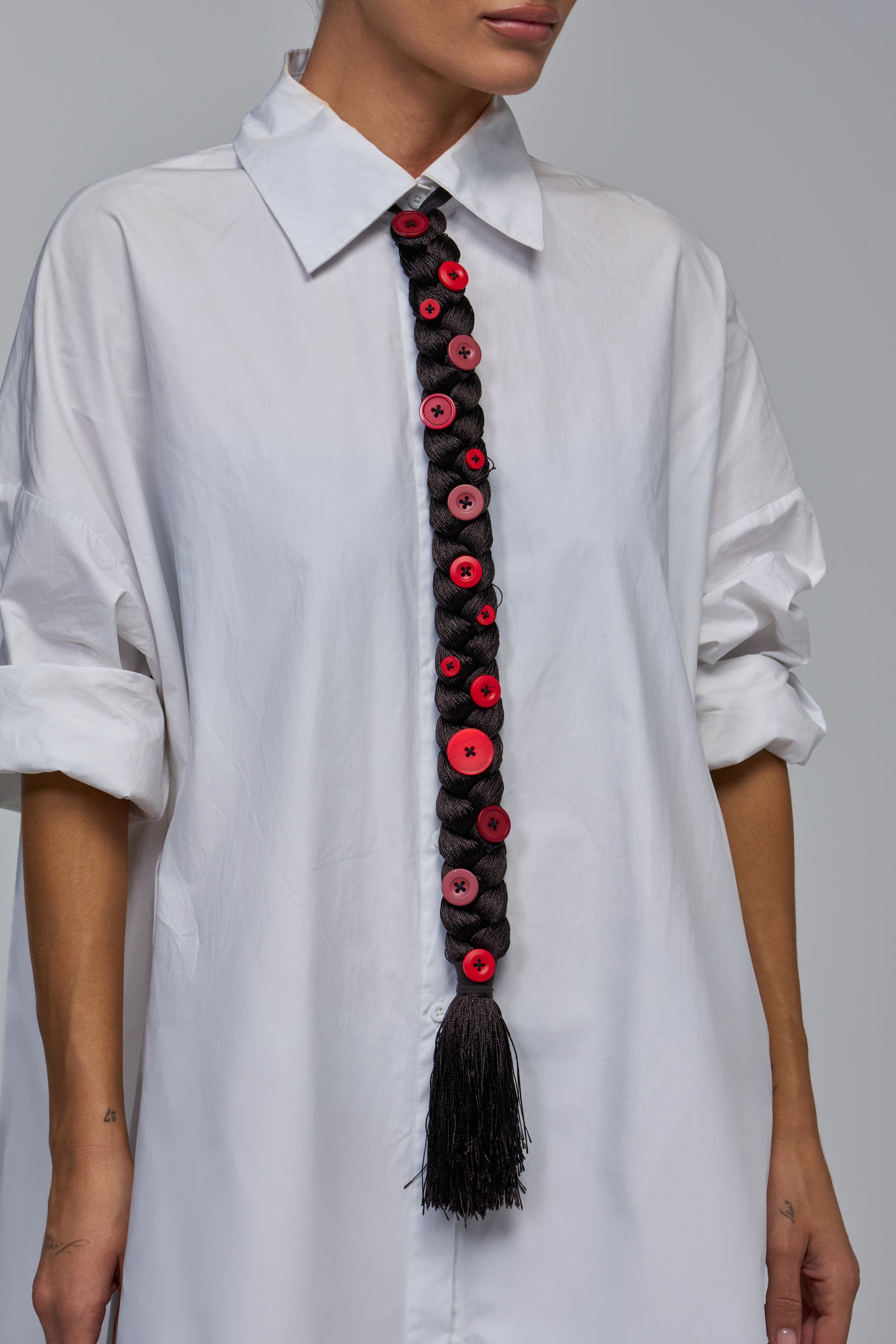 A dark brown BRAID BABY TIE embellished with a vertical row of red buttons, worn over a white collared shirt.
