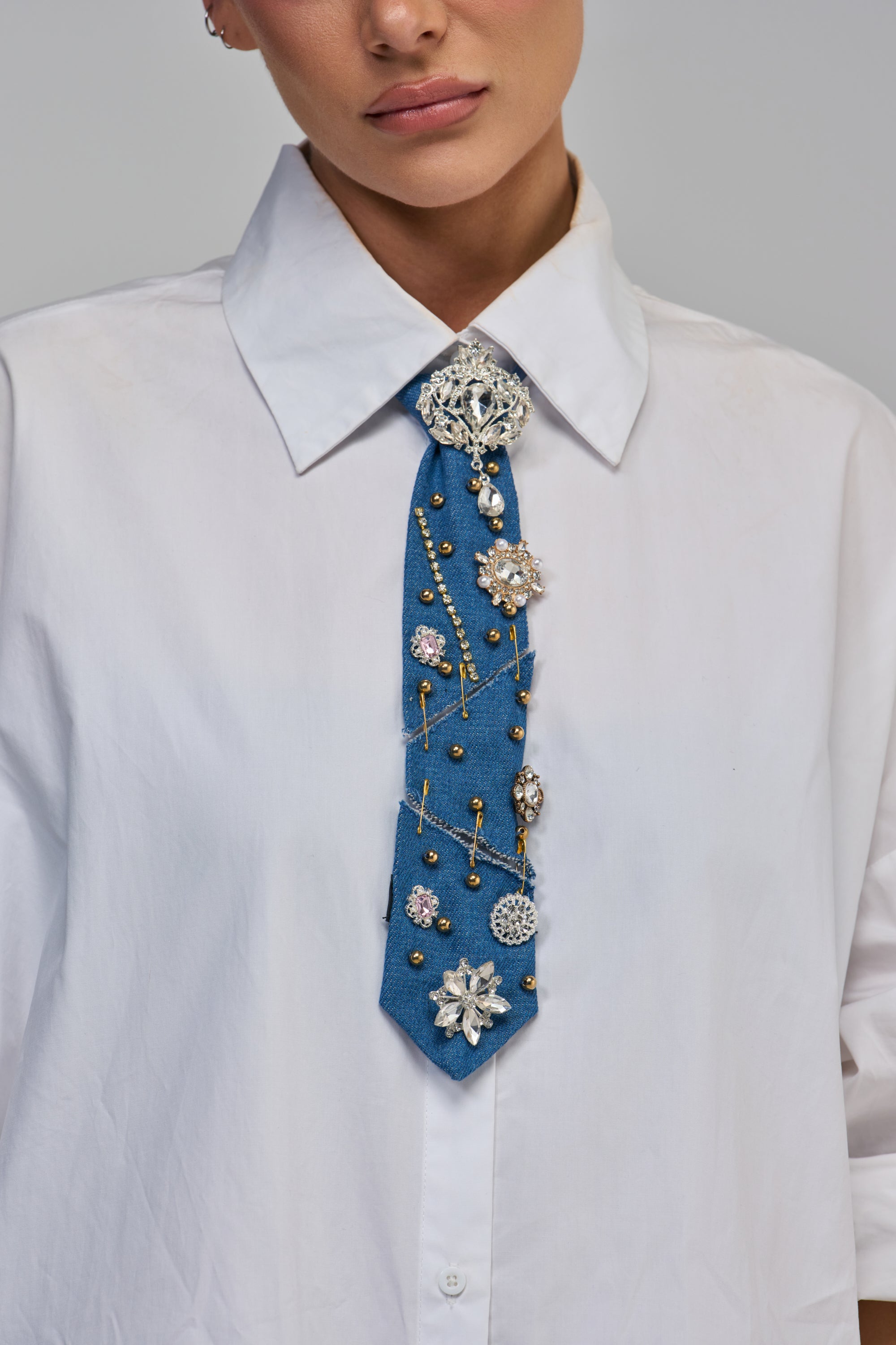 A person wearing a white collared shirt with an ALWAYS EMBELLISHED TIE, a denim tie featuring various crystals, pins, and gold studs.