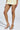 In a shiny yellow sequin mini dress, a woman stands with crossed legs, showcasing the AKIRA SACHITA SILVER AND GOLD STILETTO SANDALS featuring ankle straps, a faux leather upper, and comfort cushion insoles as she holds a small clutch against the light-colored background.