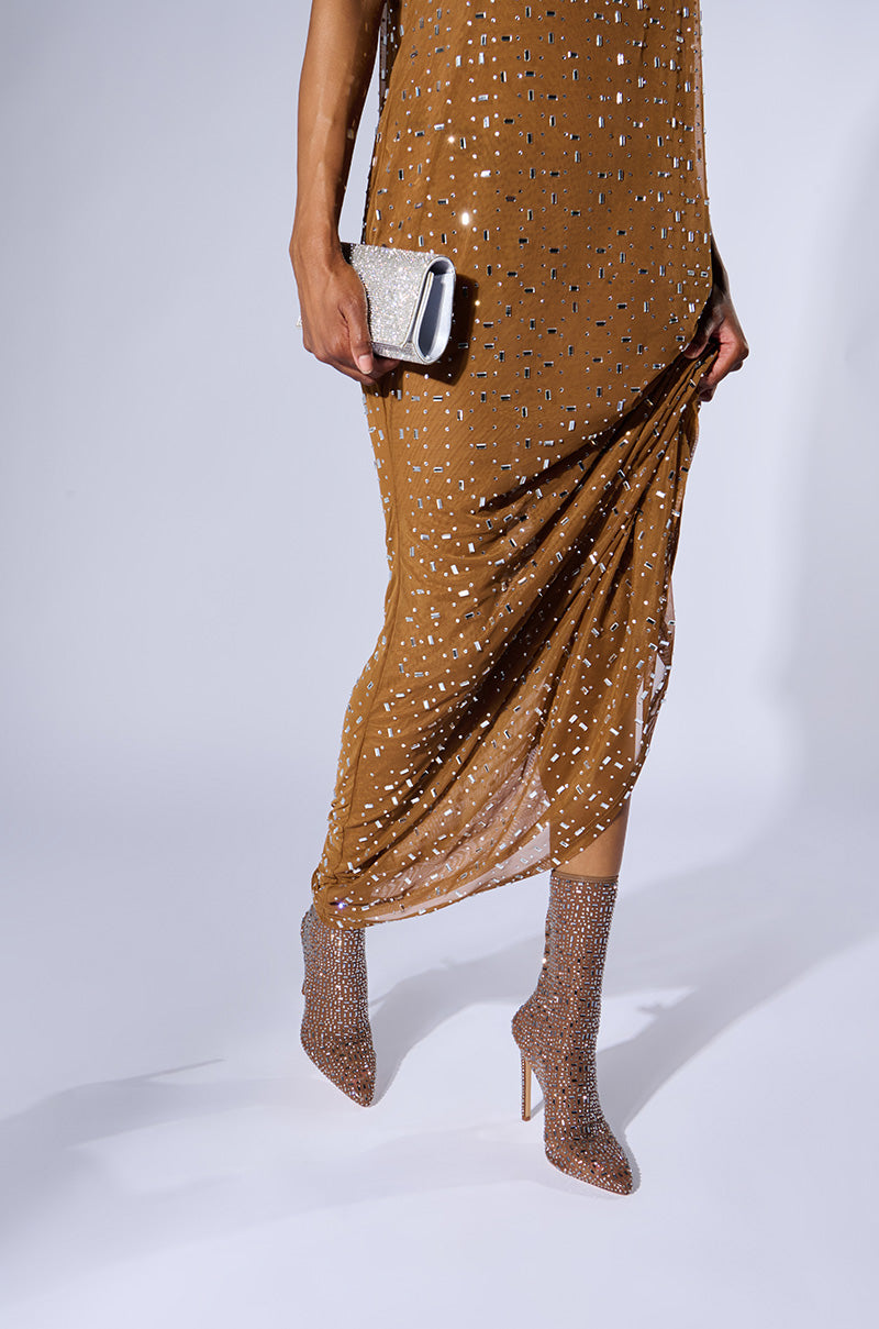 A person wearing a brown, sequined dress holds the lower part of the dress with one hand and carries a sparkly silver clutch in the other. They are also wearing AZALEA WANG SADIRA BROWN BOOTIE, which are glittery high-heeled boots. The background is plain and white.