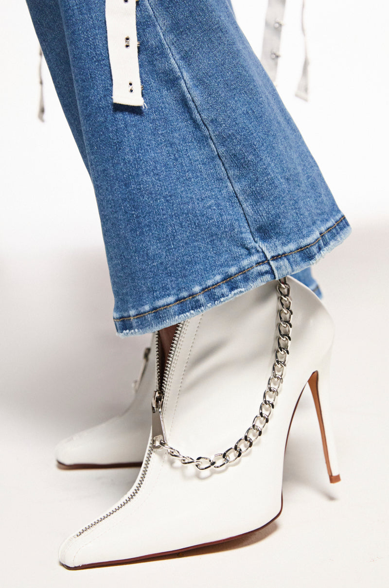 A close-up shows someone in blue denim pants and AZALEA WANG SAGITTA WHITE STILETTO PUMPS. These faux leather high-heeled boots feature pointed toes, zipper details, and a silver chain elegantly wrapping around the heel and ankle.