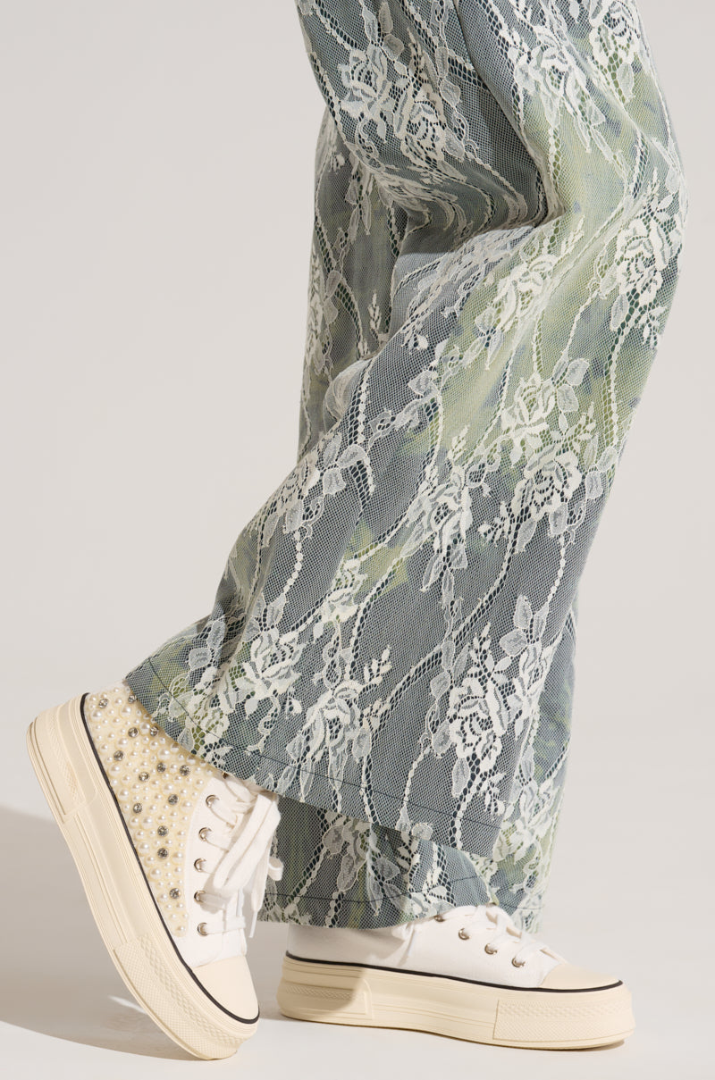 A close-up of a person's legs wearing wide-legged pants with a blue-green and white lace pattern, paired with the AZALEA WANG SAISHA PEARL WHITE SNEAKER, which features pearl and bead embellishments on the sides. The background is a plain off-white color.