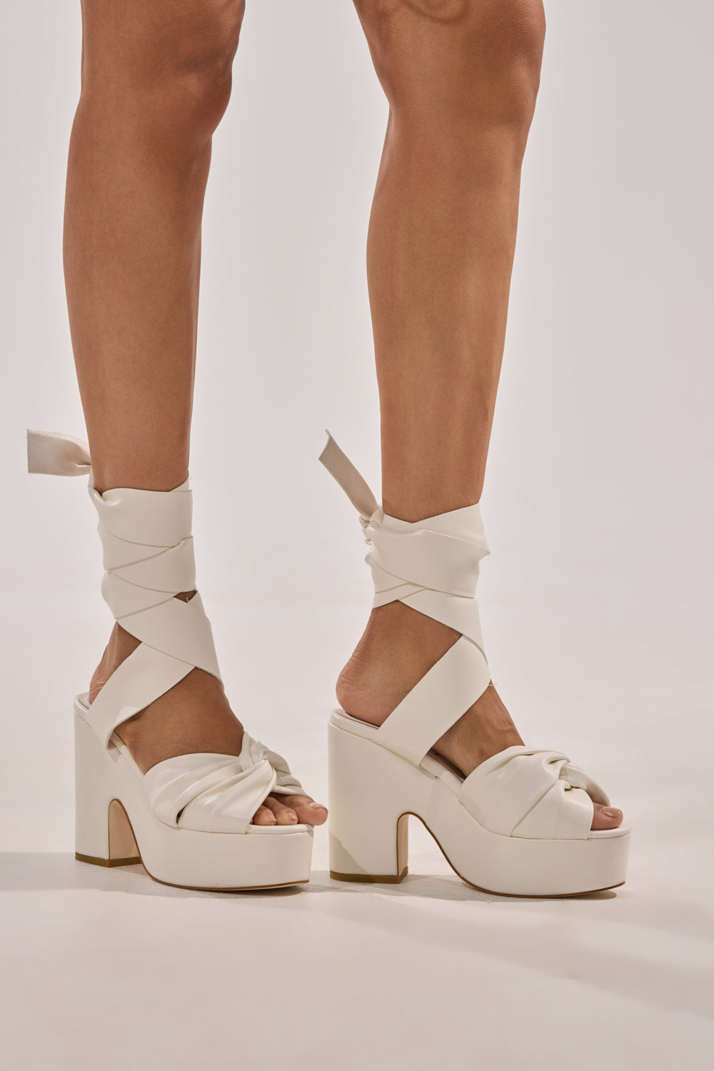A person wears the AZALEA WANG SAKI WHITE PU CHUNKY WRAP UP SANDAL, featuring chunky white faux leather and thick ankle wrap straps tied in bows, standing on a plain light background. 8