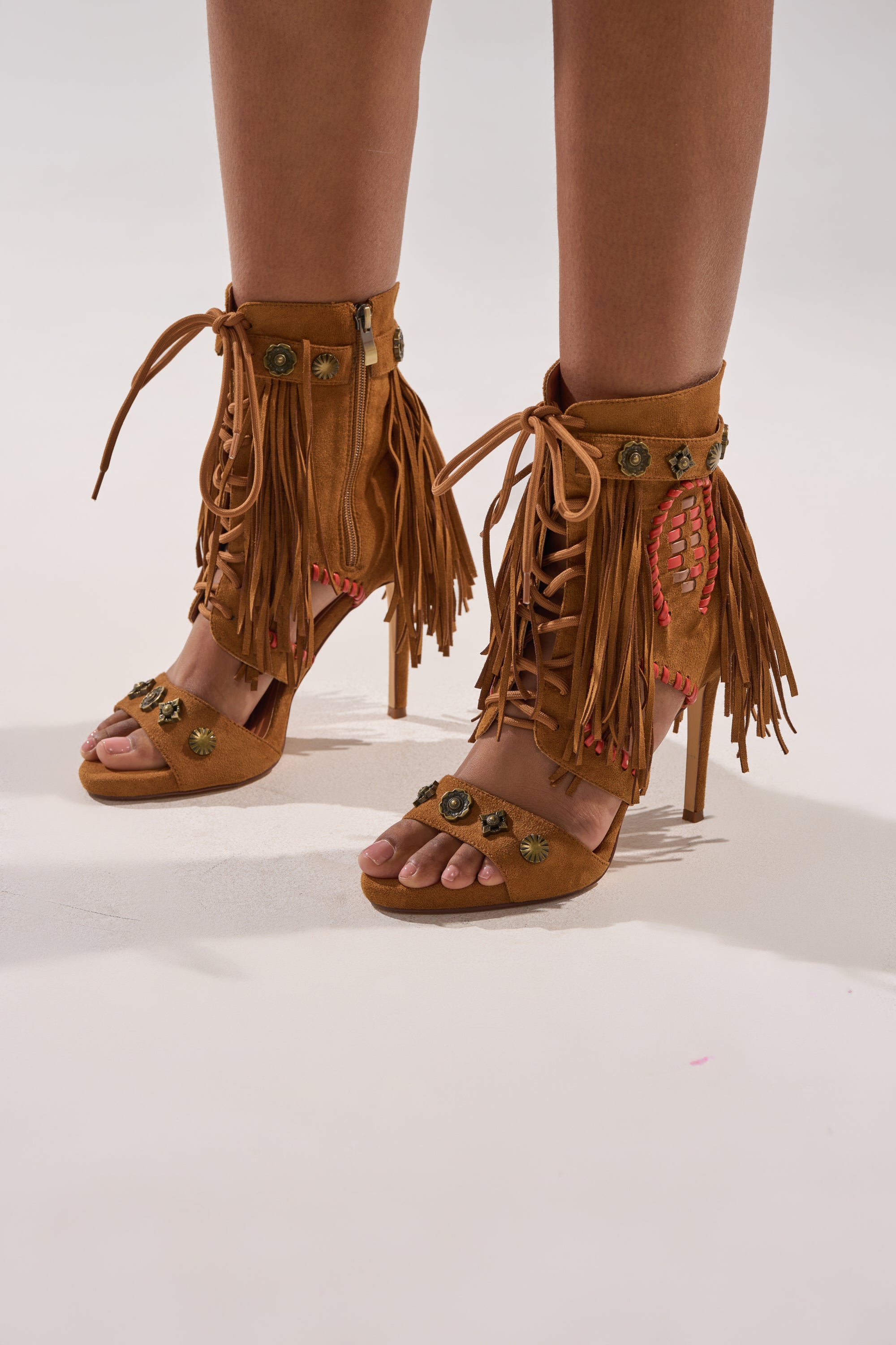 A person wears the AZALEA WANG SALGADO FRINGE GLADIATOR STILETTO SANDAL—boho-inspired brown suede, open-toe, high-heeled boots with fringe, lace-up fronts, and decorative details—while standing on a light surface.