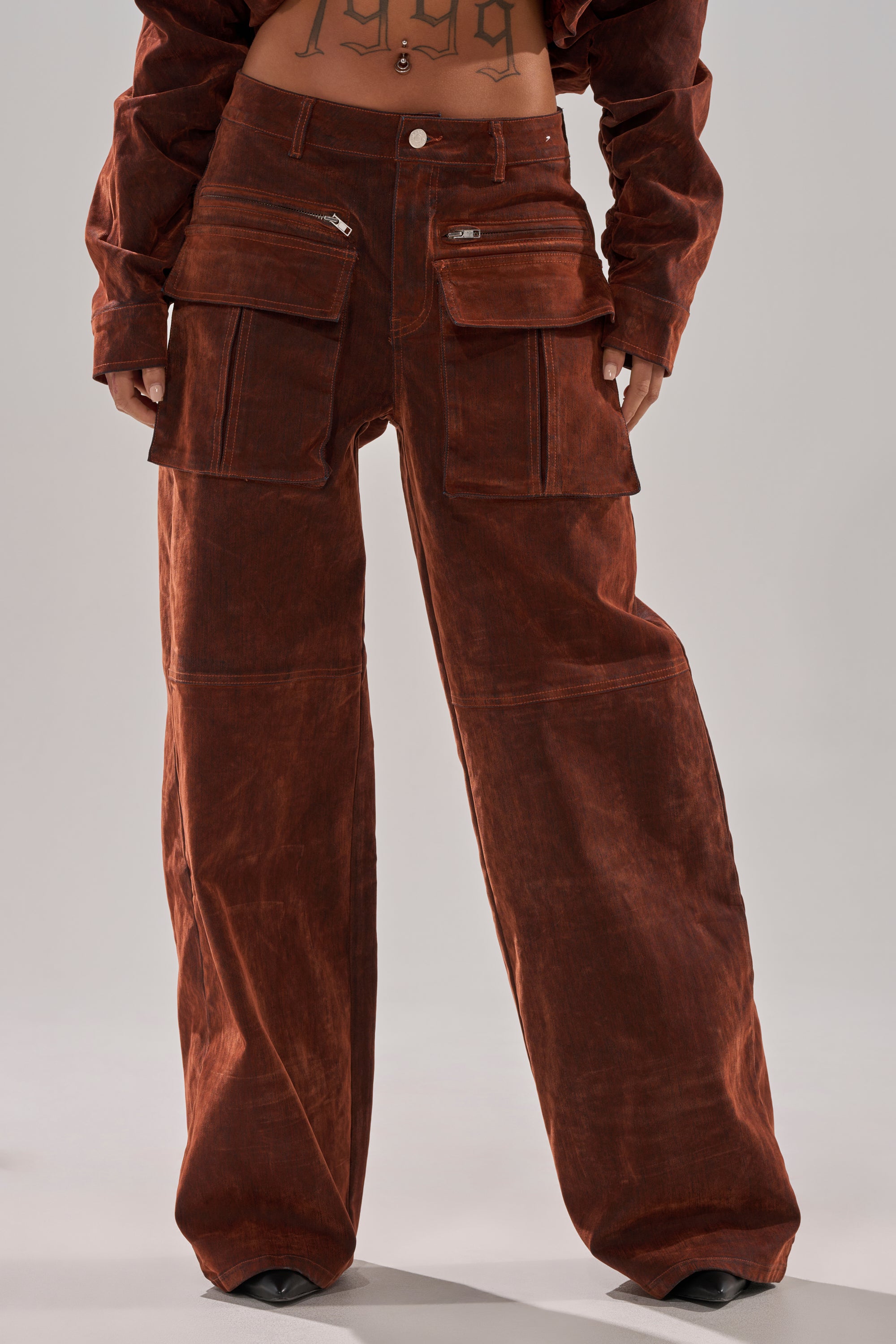 A person wears the AKIRA Label THINK ABOUT YOU ALL THE TIME VELVET DENIM CARGO TROUSER—high-waisted, wide-leg cargo pants with large front pockets and zippers—paired with a matching jacket, exposing a tattooed midriff.