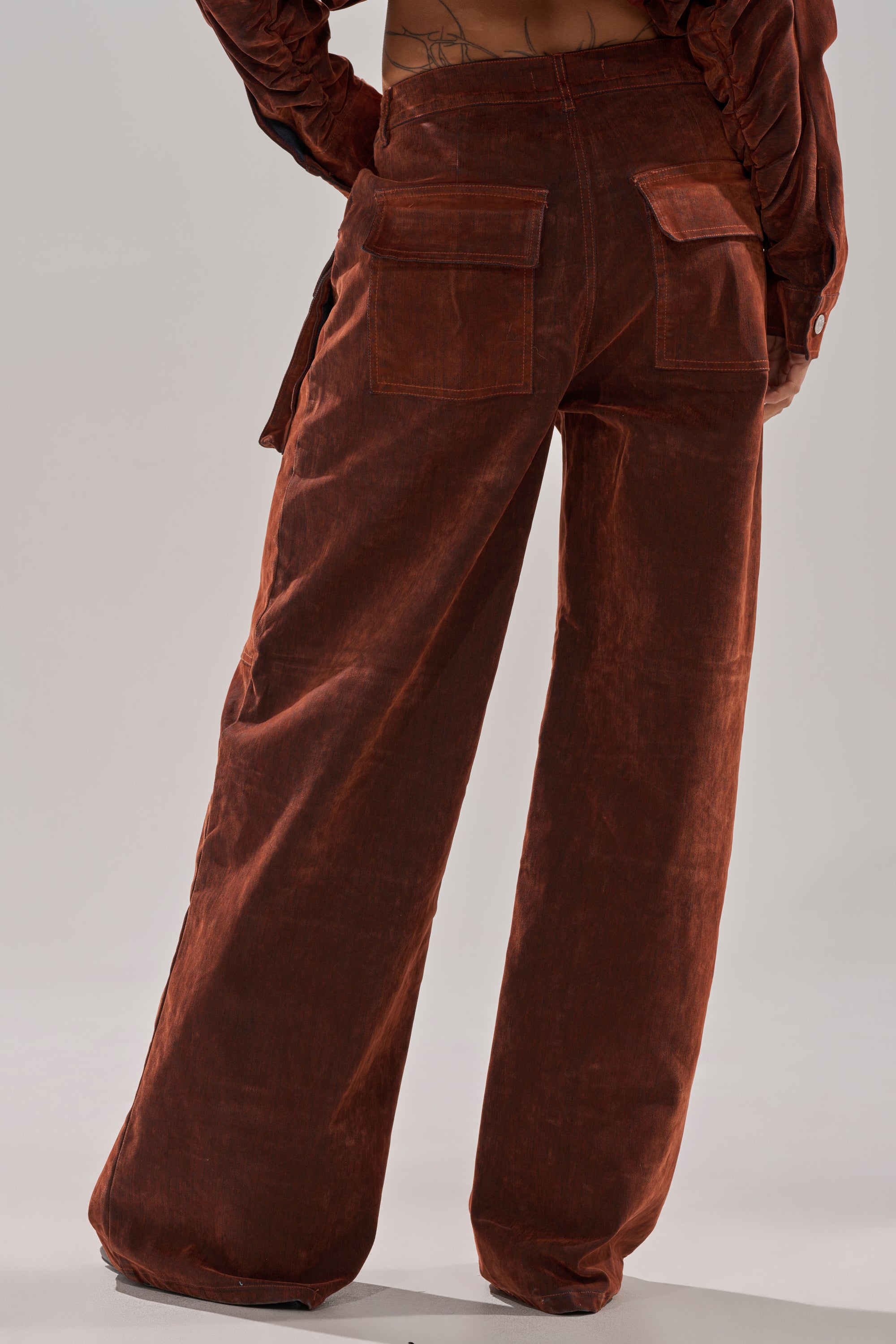 A person wearing loose-fitting, rust-colored THINK ABOUT YOU ALL THE TIME VELVET DENIM CARGO TROUSER with large back pockets stands with their back to the camera against a neutral background.