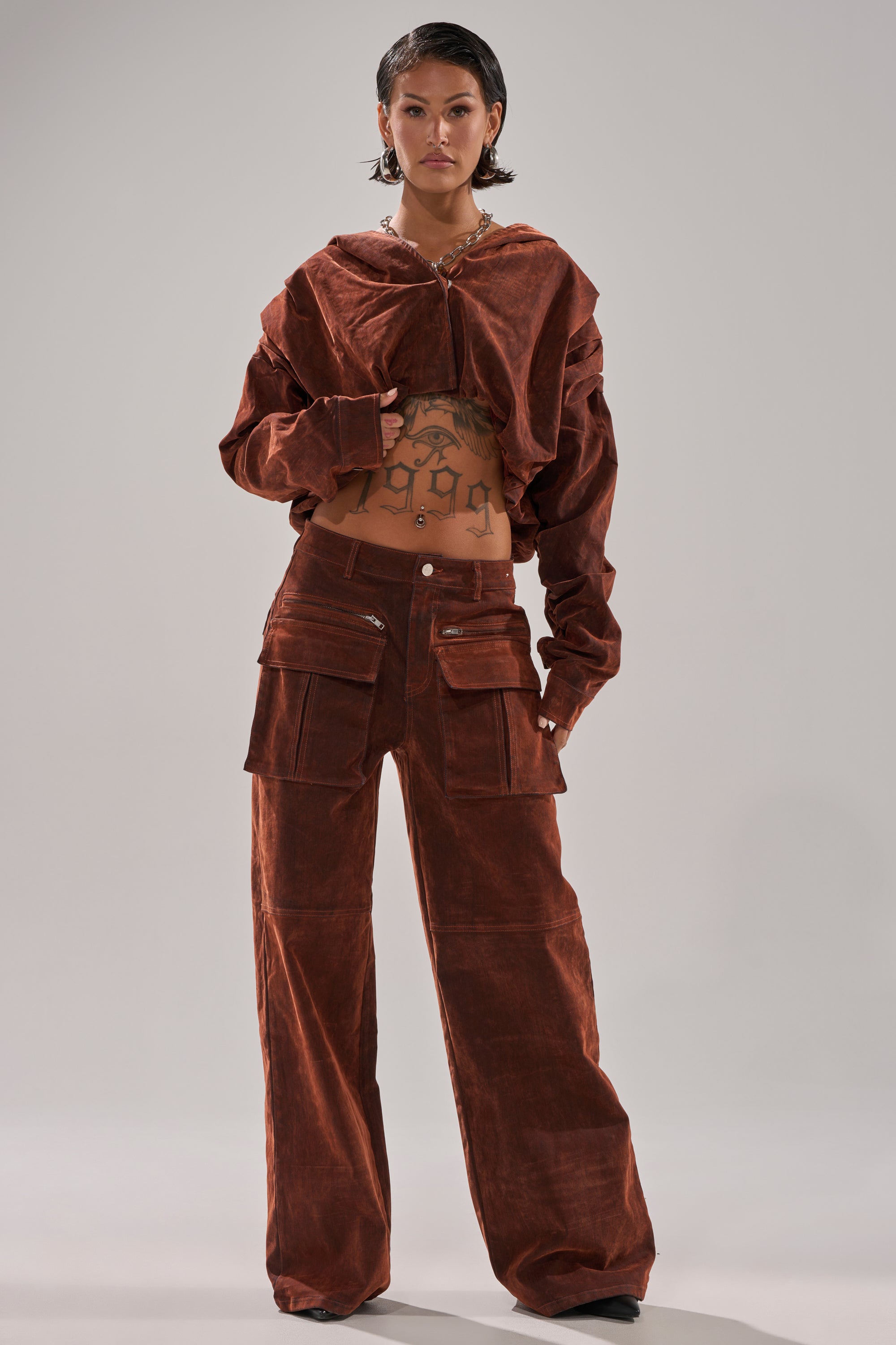 A person stands facing forward in a brown, oversized corduroy jacket and the THINK ABOUT YOU ALL THE TIME VELVET DENIM CARGO TROUSER, which features large front pockets. Their midriff with tattoos is visible; they have short hair and a confident expression.