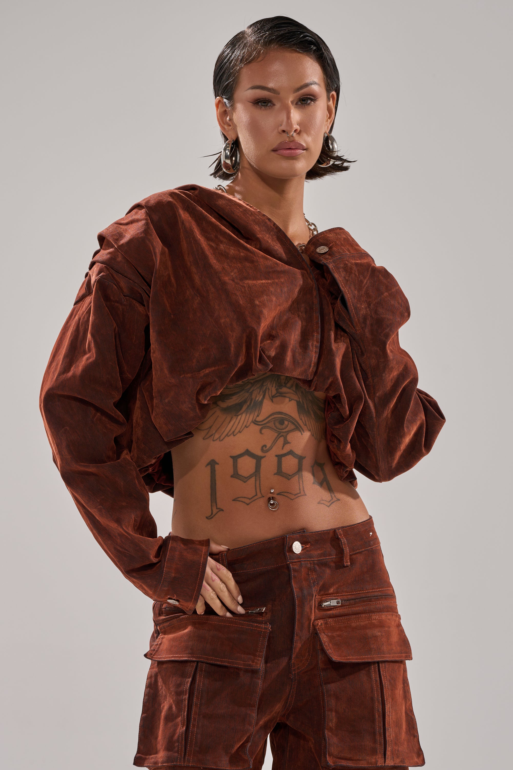 A person with short dark hair wears the THINK ABOUT YOU ALL THE TIME VELVET DENIM LONG SLEEVE TOP, revealing a large "1995" tattoo with an eye design on their stomach, and poses confidently against a plain background.