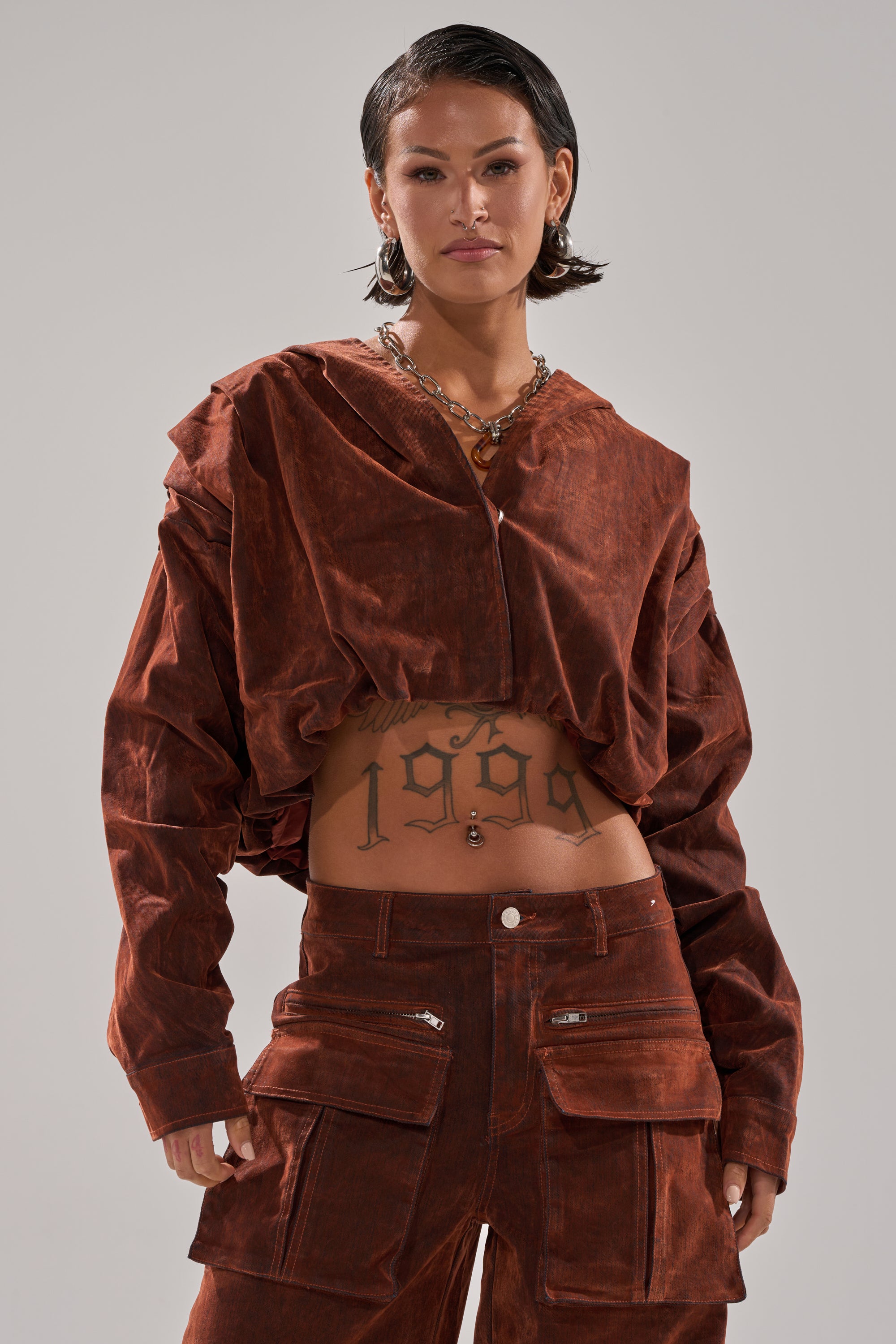 A person with short dark hair wears the THINK ABOUT YOU ALL THE TIME VELVET DENIM LONG SLEEVE TOP in rust, paired with matching pants. Their "1998" tattoo and layered necklaces complete the confidently striking look.