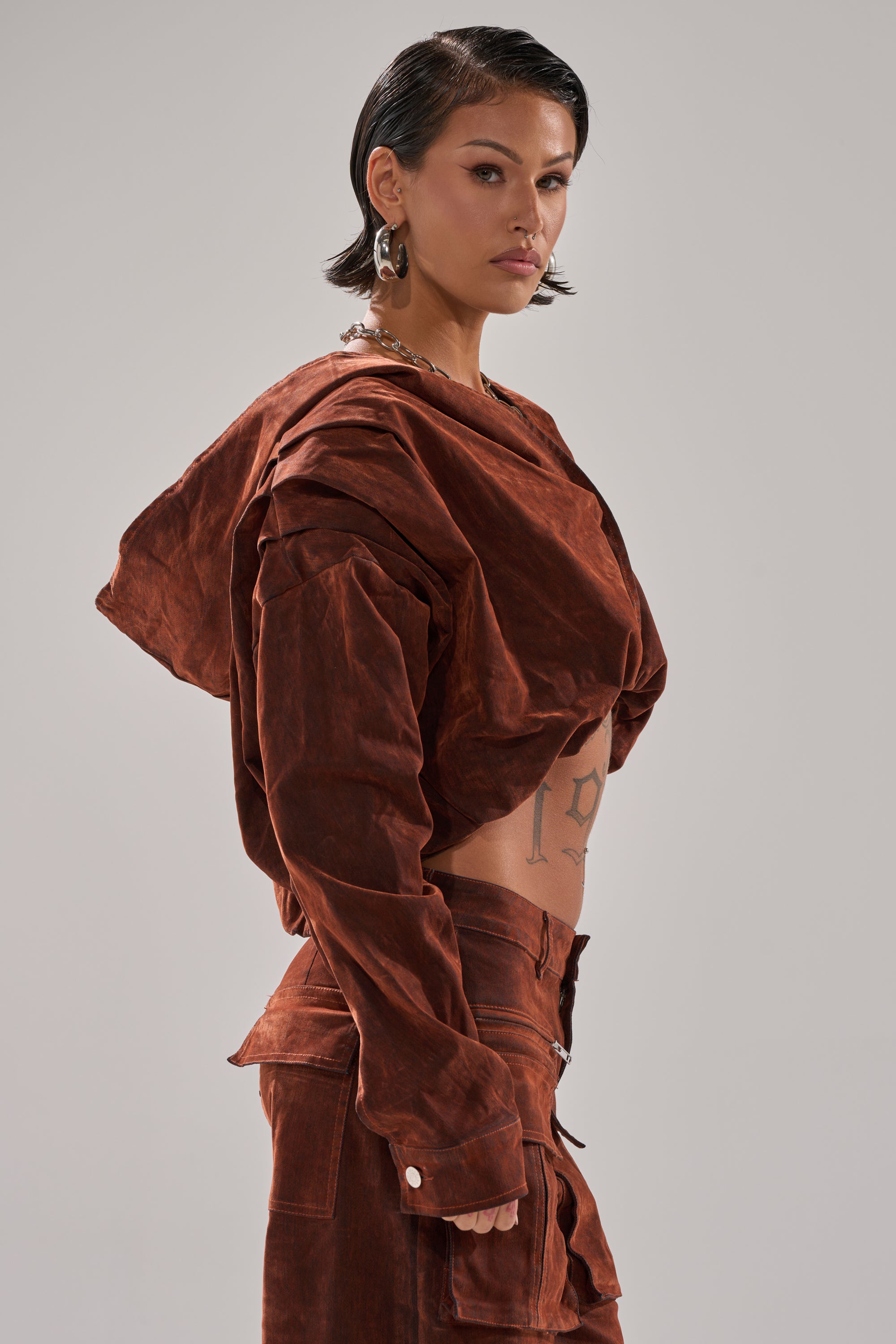 A person with short dark hair wears the THINK ABOUT YOU ALL THE TIME VELVET DENIM LONG SLEEVE TOP, hoop earrings, and shows tattoos on their midriff while posing confidently against a neutral background.