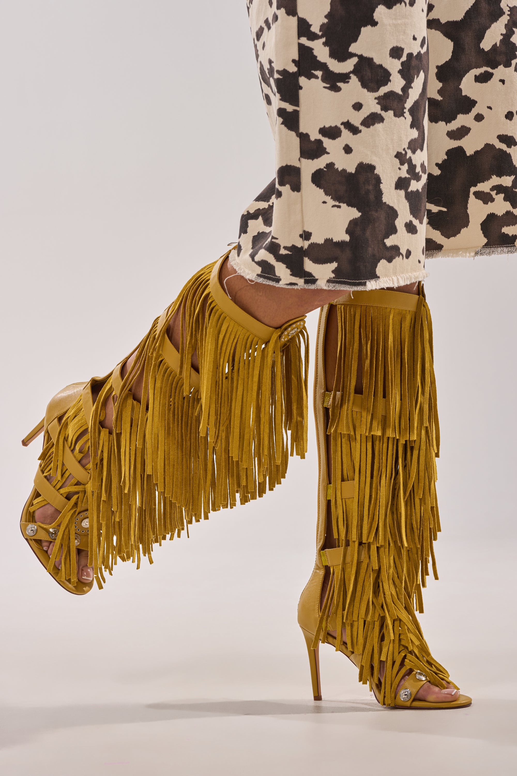 A person wears the AZALEA WANG SAMEYANA Mustard Fringe and Metal Stiletto Gladiator Sandals with knee-high fringe, paired with a black and white cow-print skirt or dress. 0