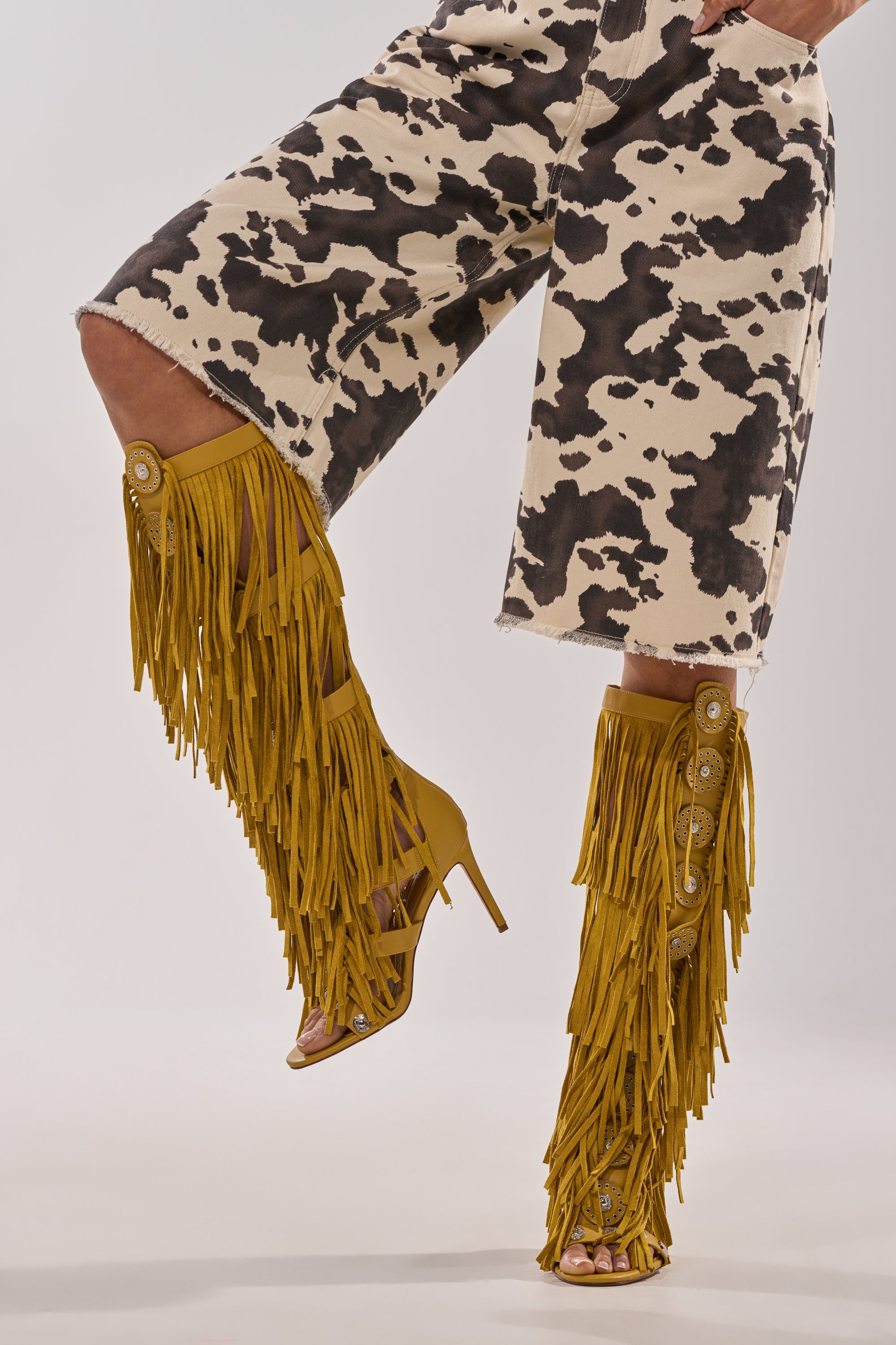 Wearing the AZALEA WANG SAMEYANA MUSTARD FRINGE AND METAL STILETTO GLADIATOR SANDAL with yellow and black-and-white cow print knee-length shorts, one leg bent and the other straight against a plain background. 1