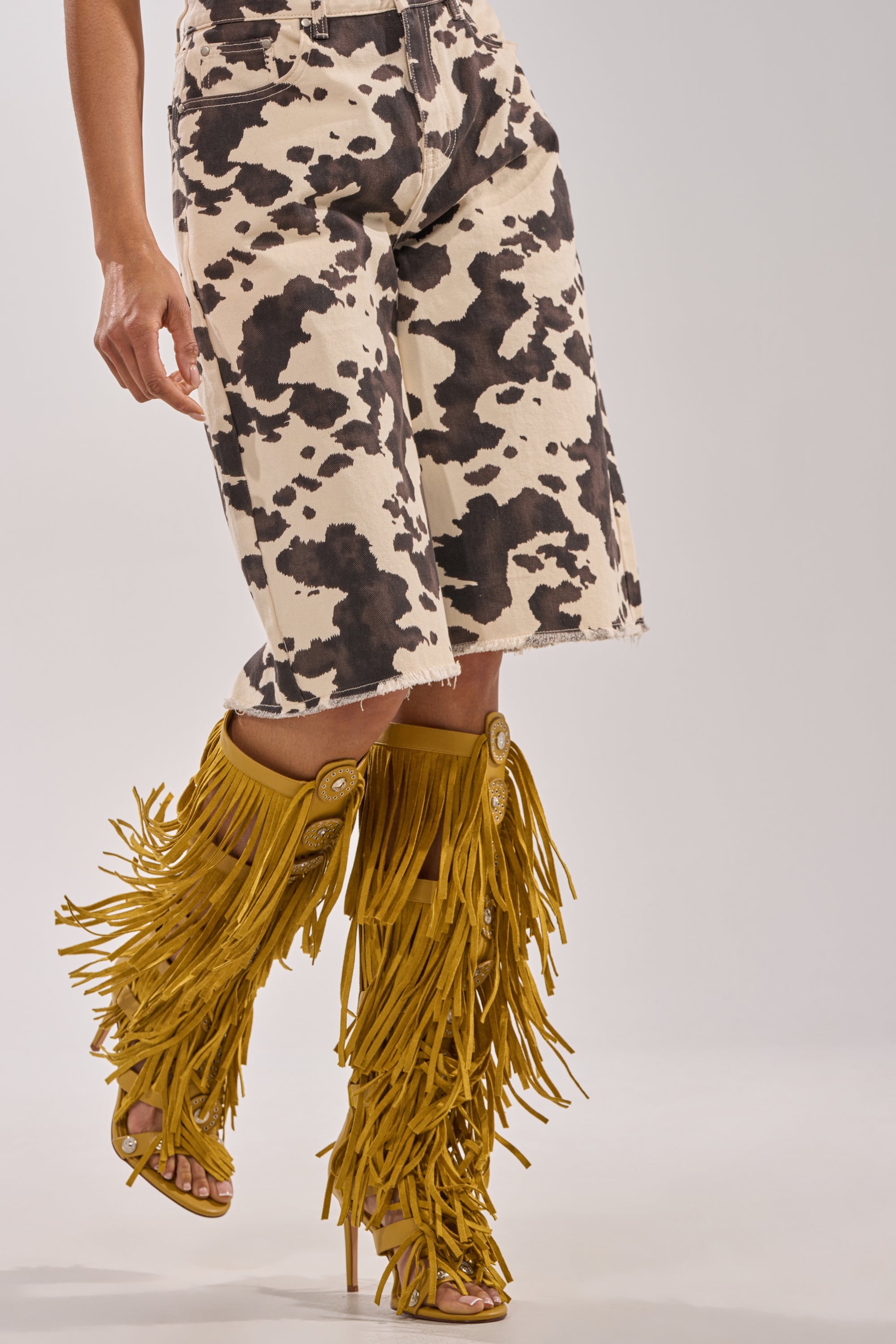 A person wears knee-length, black and white cow-print shorts and AZALEA WANG SAMEYANA mustard fringe and metal stiletto gladiator sandals. The background is plain, light gray. 2