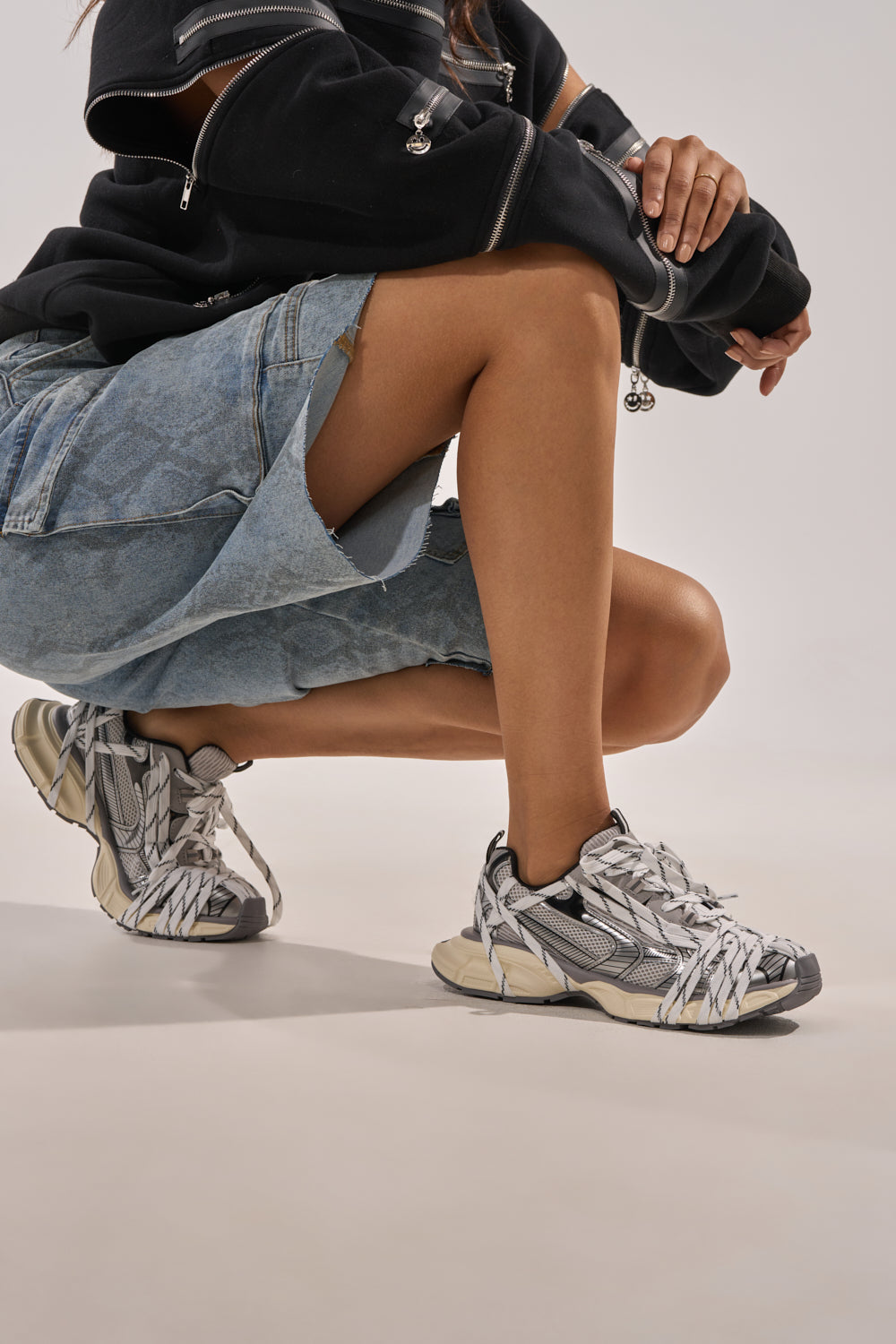 A person squats with one knee bent, wearing a black hoodie, a denim skirt with a front slit, and AZALEA WANG SAMURAI GREY MULTI CHUNKY SNEAKERS. The background is plain and light-colored. 0