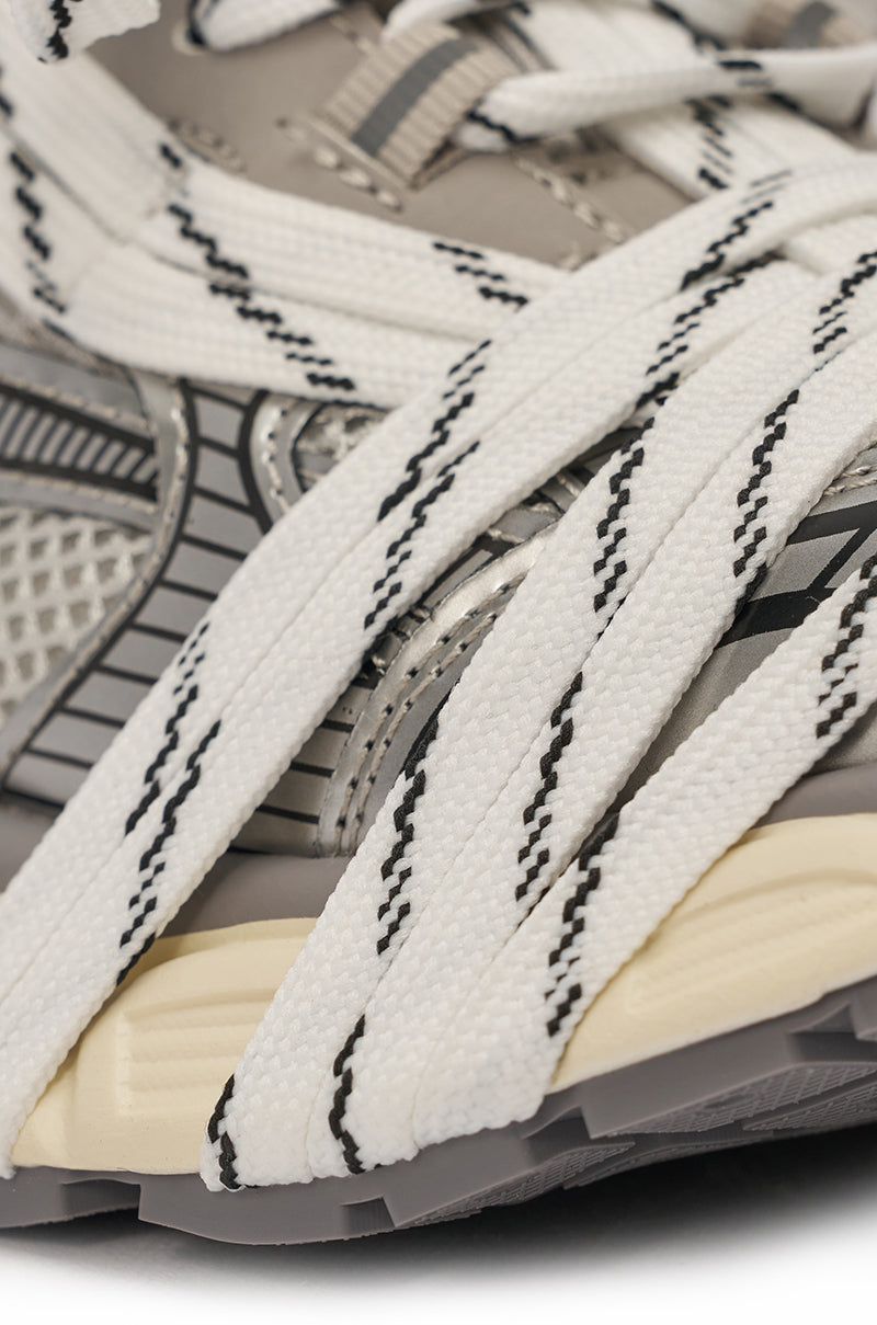 Close-up of the AZALEA WANG SAMURAI GREY MULTI CHUNKY SNEAKER, featuring wide white laces with black zigzags, crisscrossed over a light gray mesh and synthetic upper with metallic accents and a beige midsole. 6